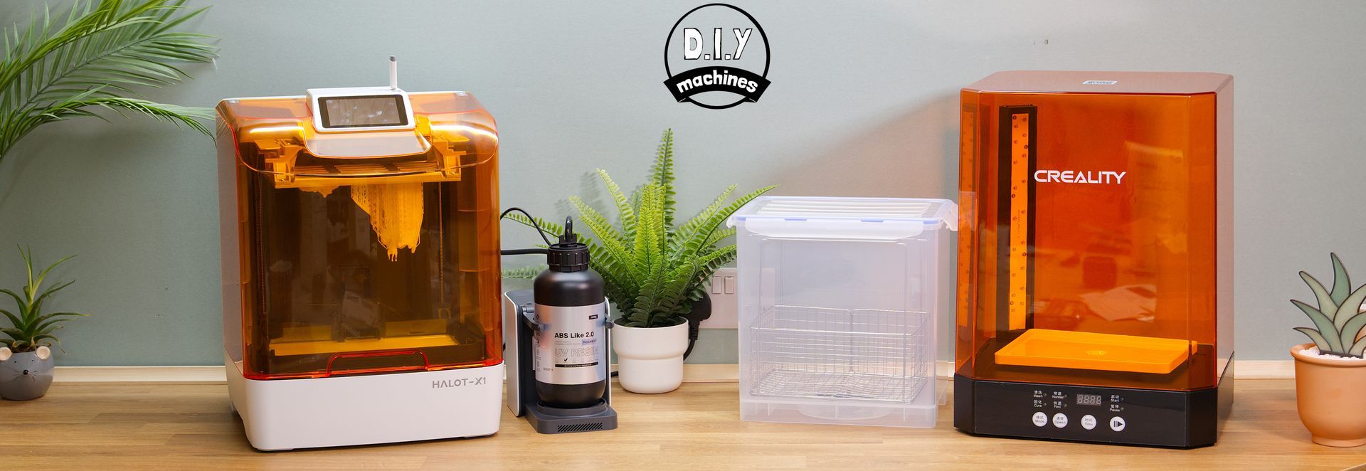 Two orange-tinted 3D resin printers, a clear wash container, and small potted plants are arranged on a wooden surface.