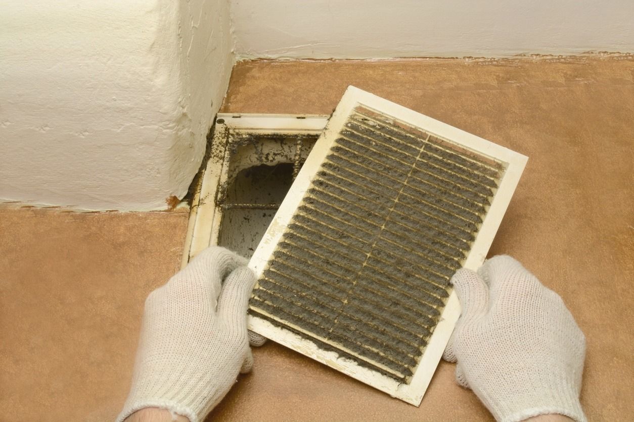 Gloved hands holding a dirty air vent cover. Brown wall, white ceiling. Dust and debris visible.