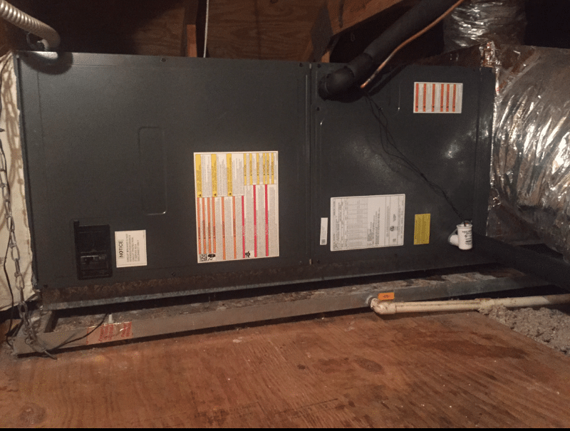 HVAC unit in an attic space with ductwork and a wooden floor. The unit is dark and has several stickers on it.