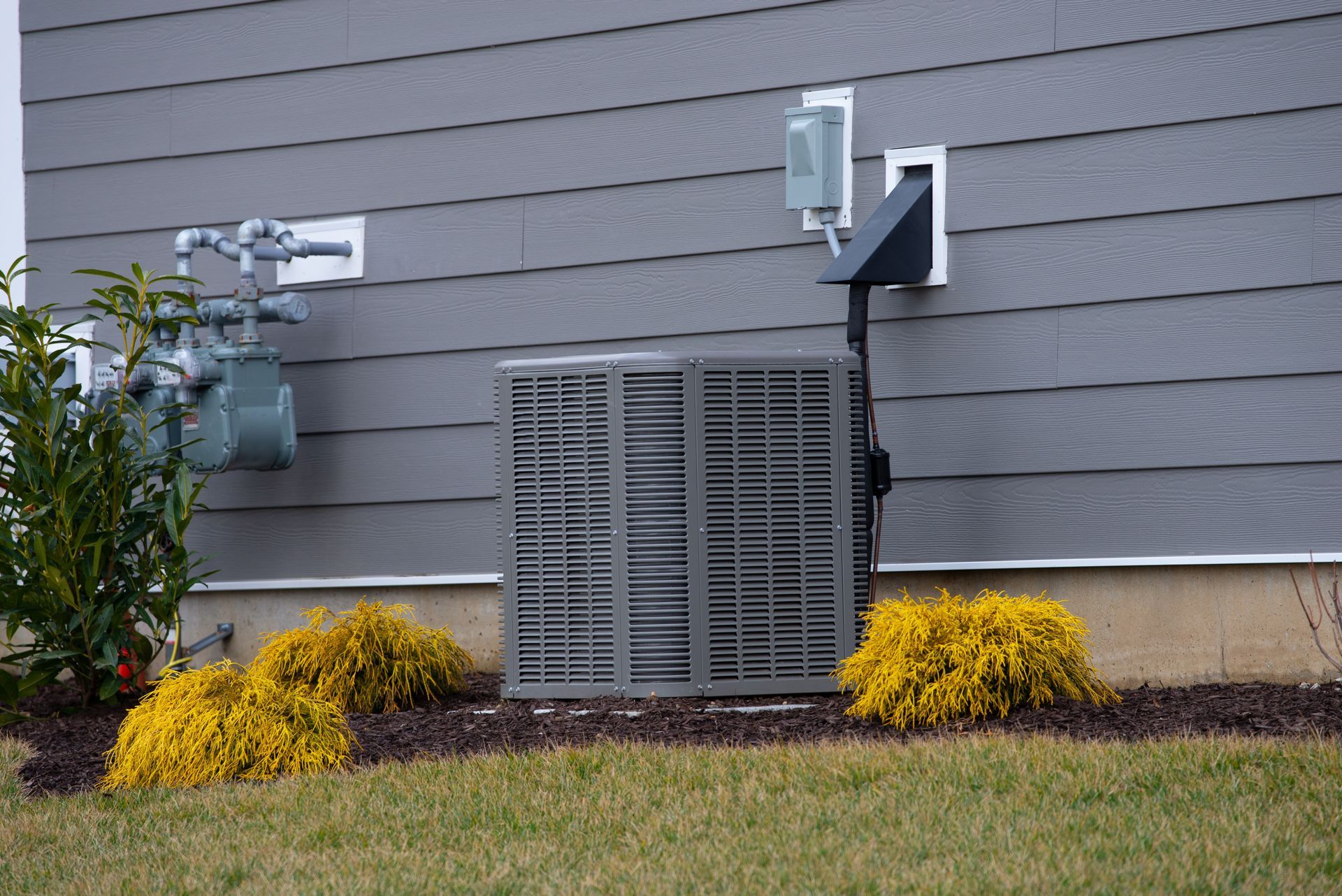 Exterior of a house with gray siding, an air conditioning unit, gas meter, and landscaping.