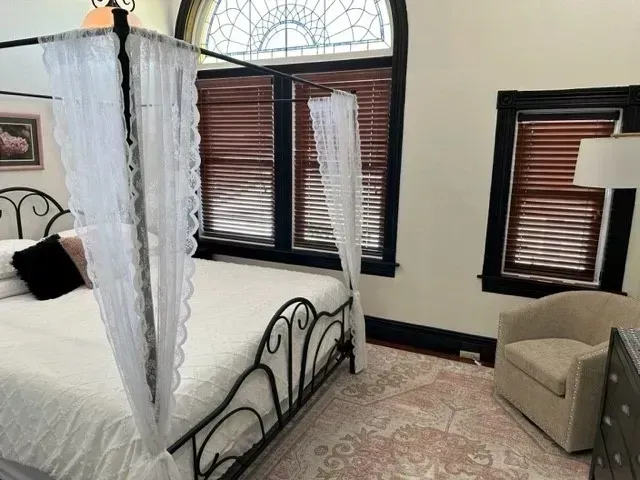 Bedroom with a four-poster bed, beige armchair, and dark-trimmed windows with blinds and sheer curtains.