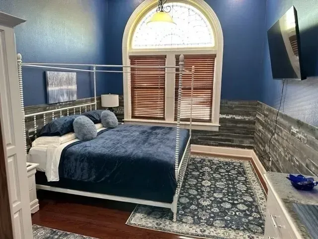 Blue bedroom with bed, arched window, patterned rug, and wall-mounted TV