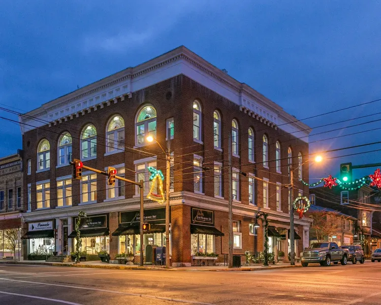 Historic brick corner building at dusk with lit windows, streetlights, and traffic in a city intersection