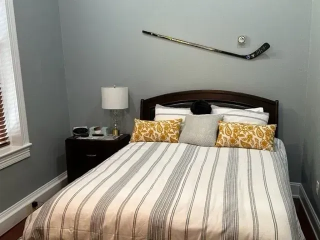 Neatly made bed with striped bedding and hockey sticks mounted above in a gray bedroom.