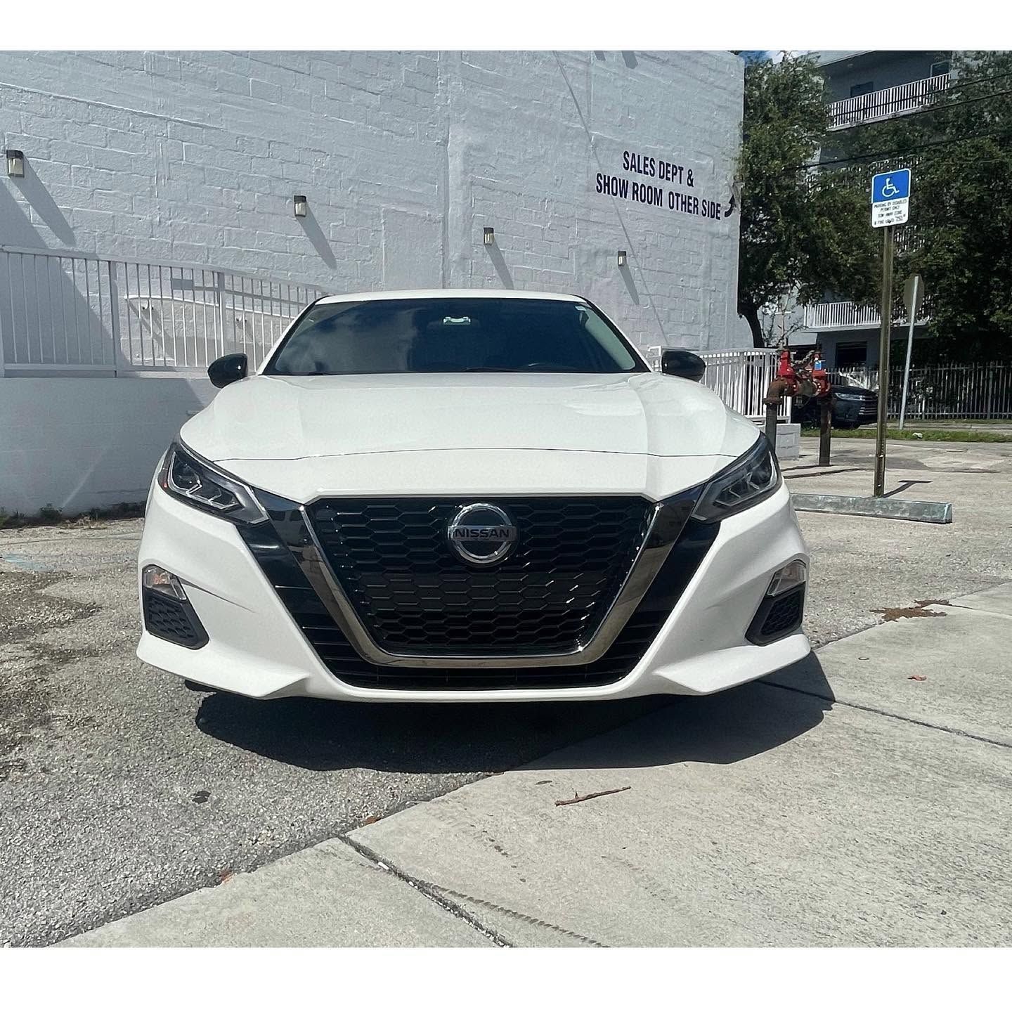 White Nissan sedan parked outside a building on a sunny street, front view