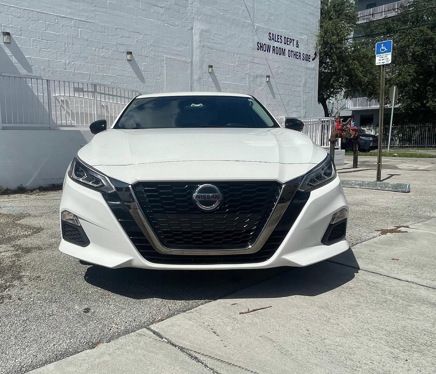 White Nissan sedan parked on a sidewalk beside a building, under bright daylight.