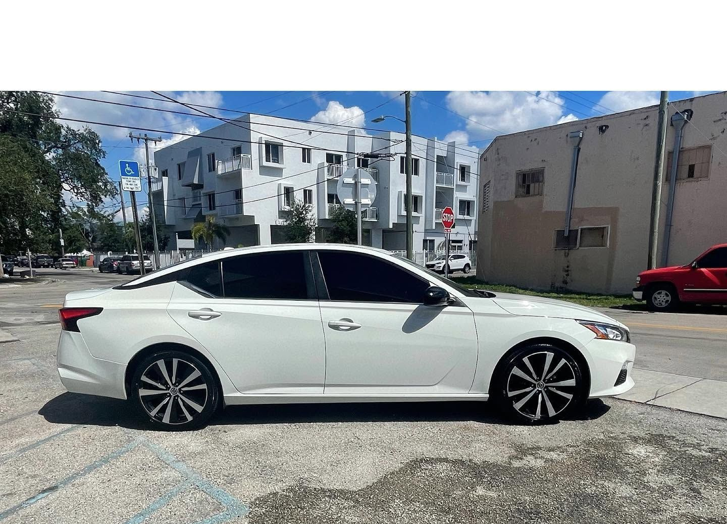 White sedan parked on a sunny street beside buildings and a red car