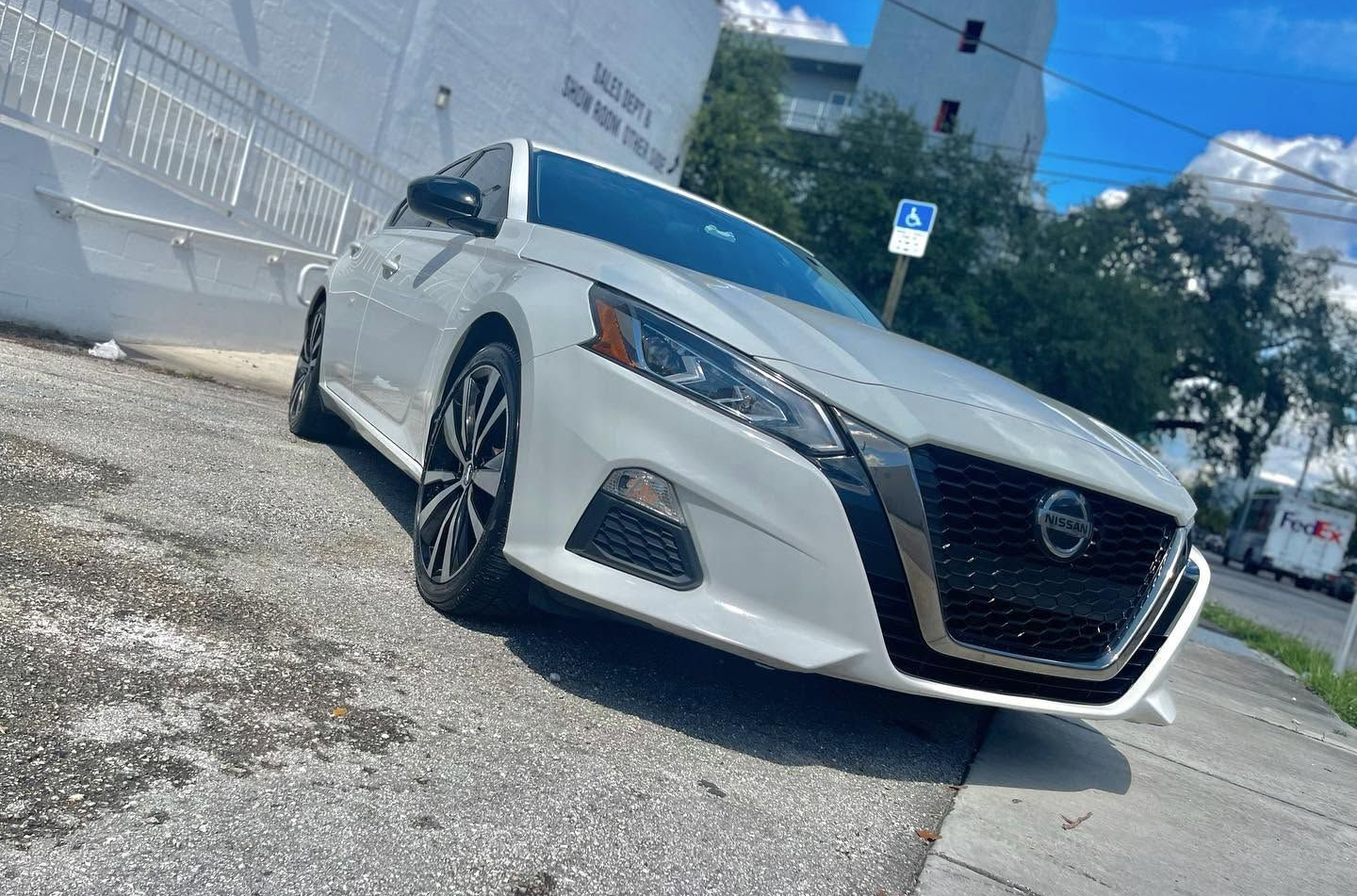 White Nissan sedan parked at an angle on a sunlit street beside a concrete wall and trees
