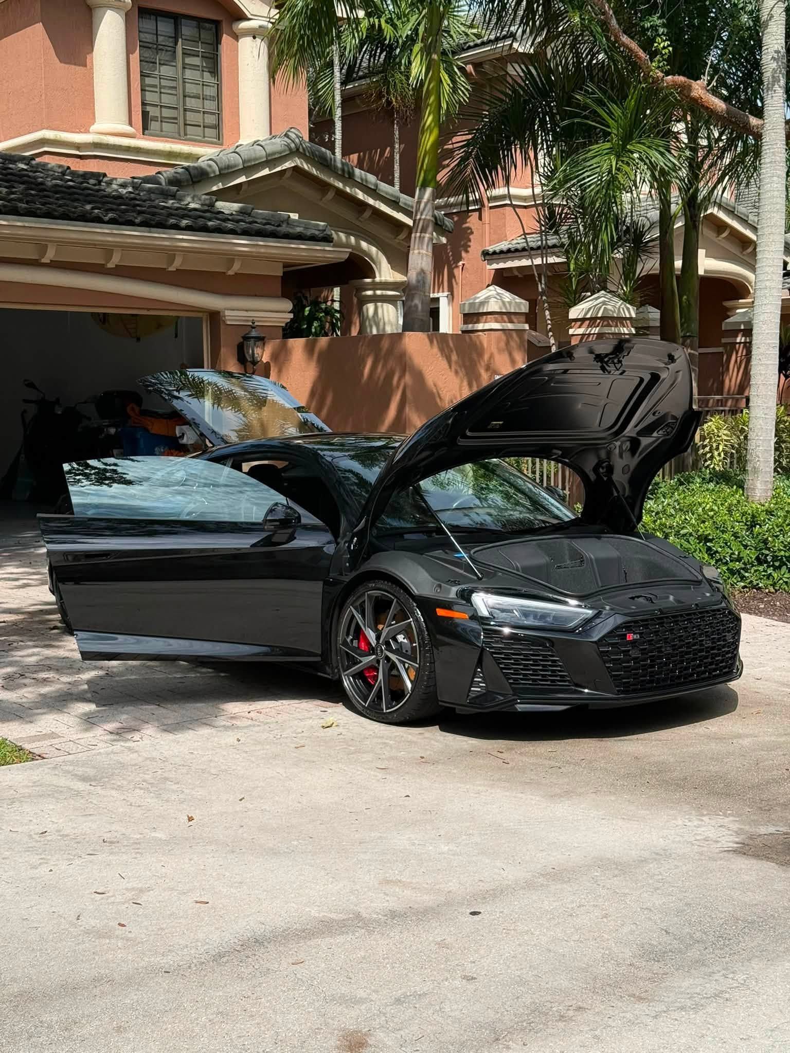 Black sports sedan with hood and driver door open, parked on a tree-lined residential street.