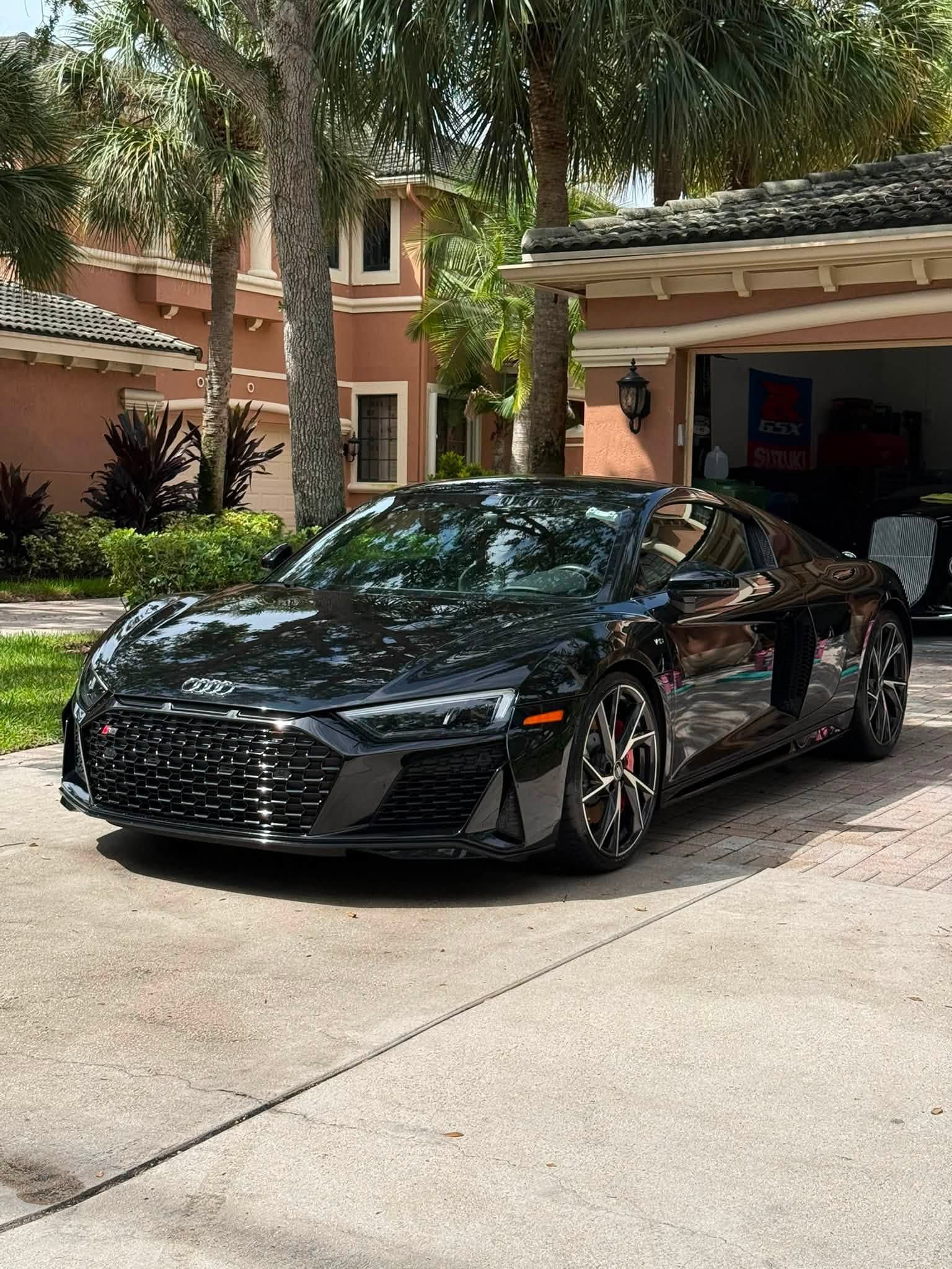 Black sports car parked in a driveway beside a house and garage, with trees in the background