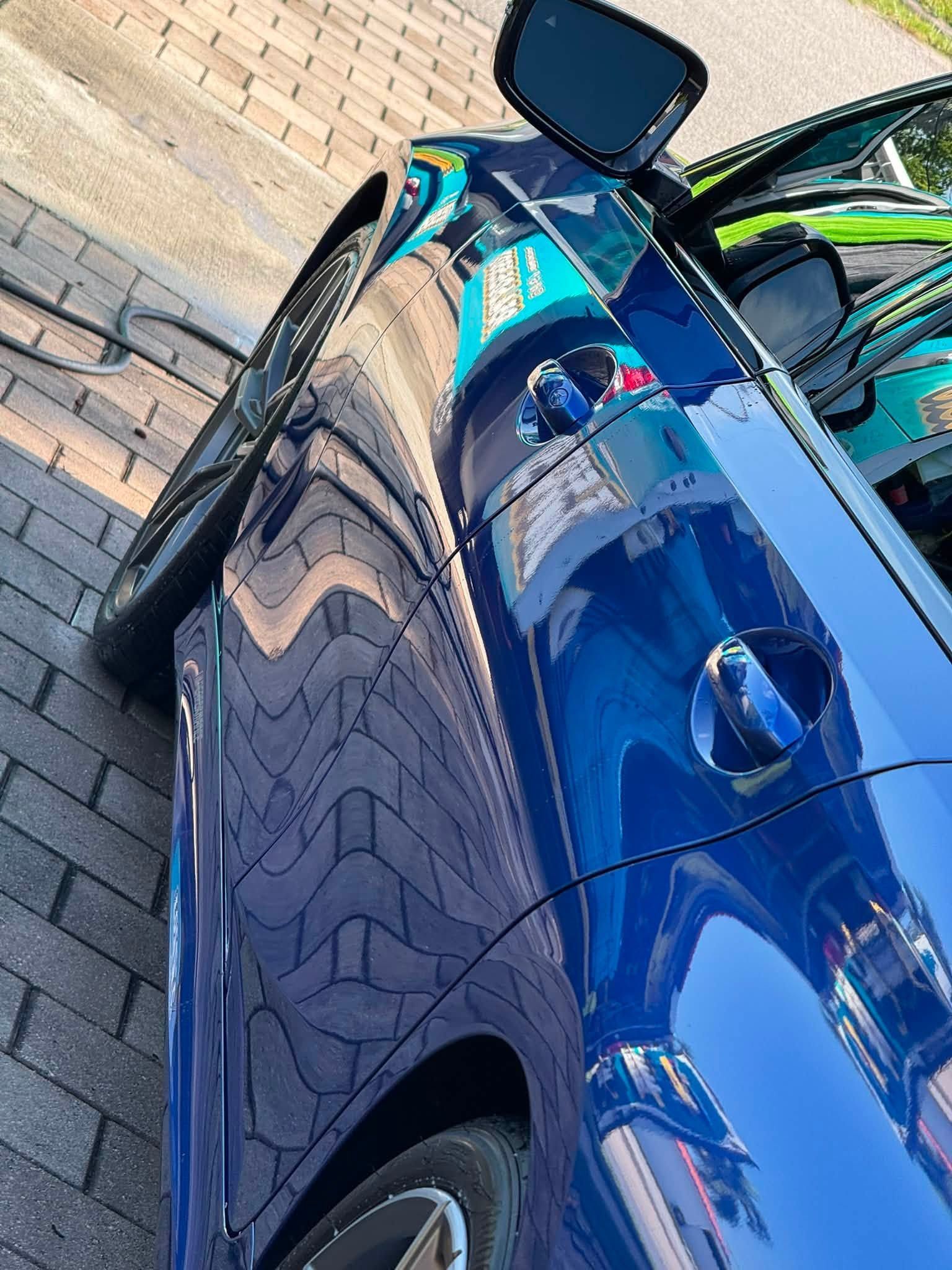 Blue car parked beside a brick wall, shown from the side with a glossy reflection.