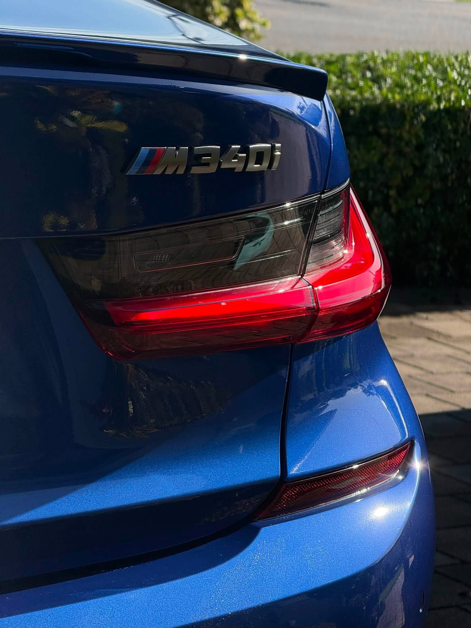 Rear of a blue BMW M340i with illuminated red taillights parked on a driveway