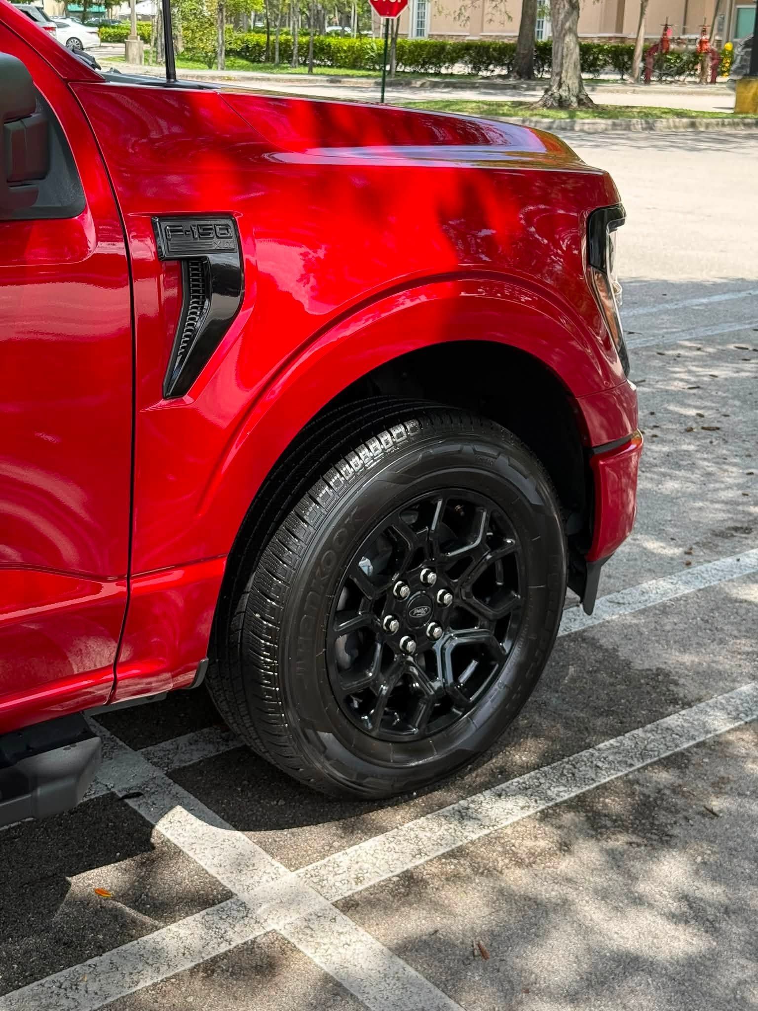 Red pickup truck parked on a sunny street, showing the front side and black wheel.