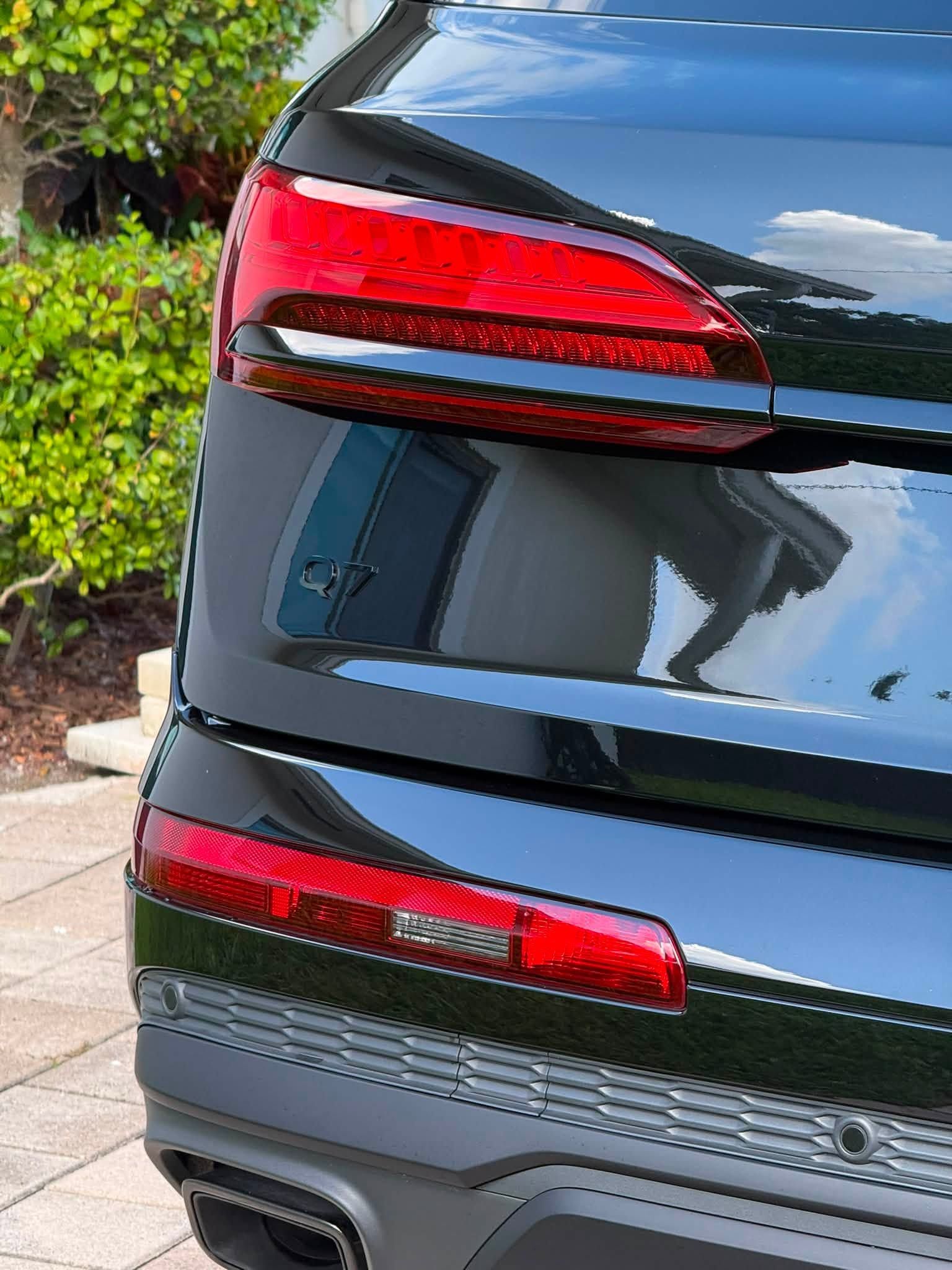 Rear of a black sedan with red taillights parked near greenery and curb paving.