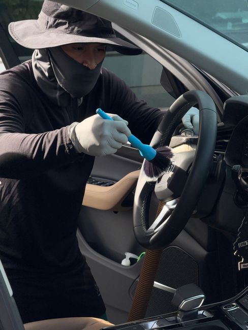Person in gloves cleaning a car steering wheel with a brush from the open driver’s side door