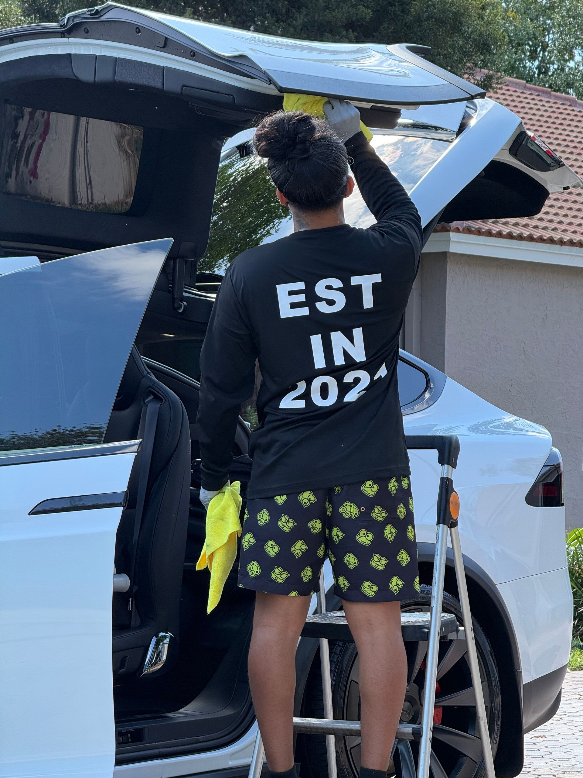 Person standing by an open car trunk, lifting a bag, wearing a black “EST IN 202” shirt and patterned shorts