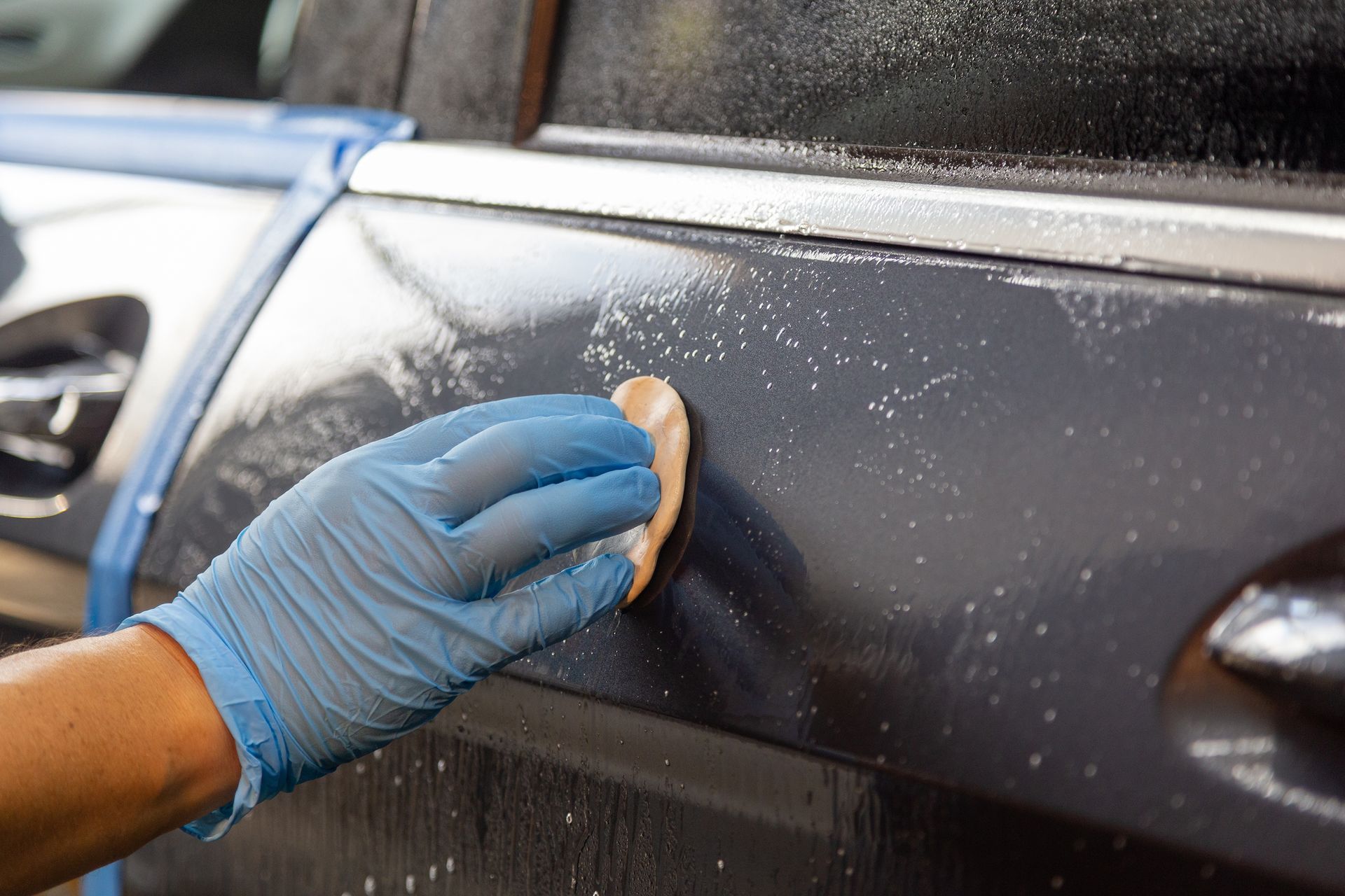 Gloved hand cleaning a black car door with a sponge during car detailing
