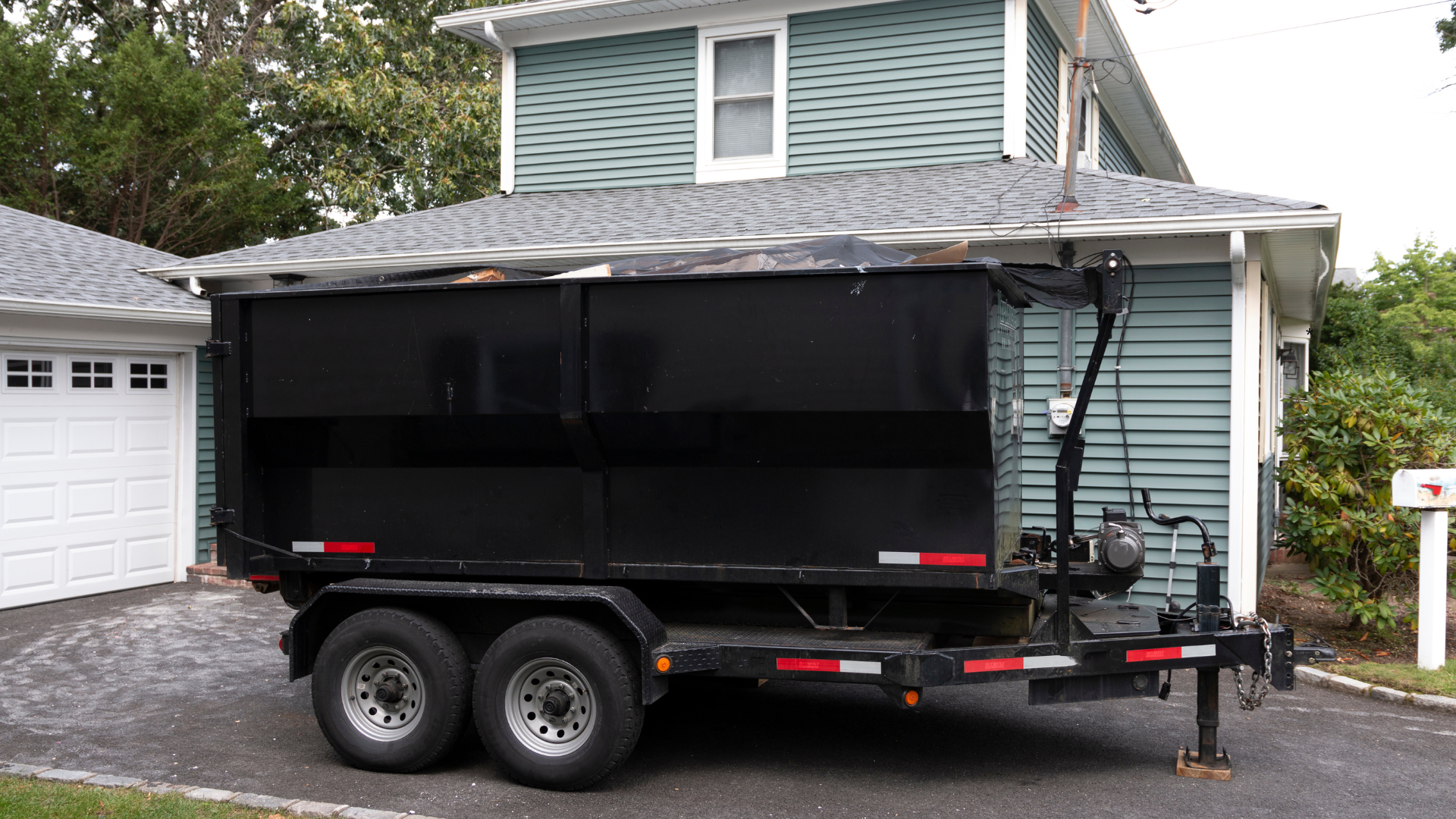 Black dumpster trailer parked in front of a house, with a white garage door on the left and a mailbox on the right.