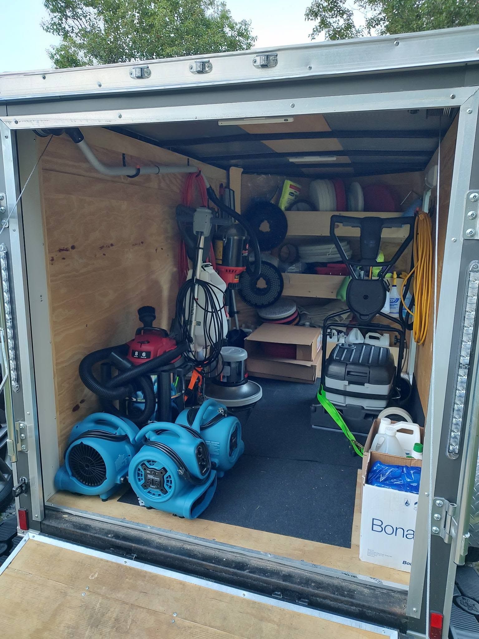 Open trailer packed with tools, blue hoses, shelves, a folded chair, and boxes inside wooden walls.