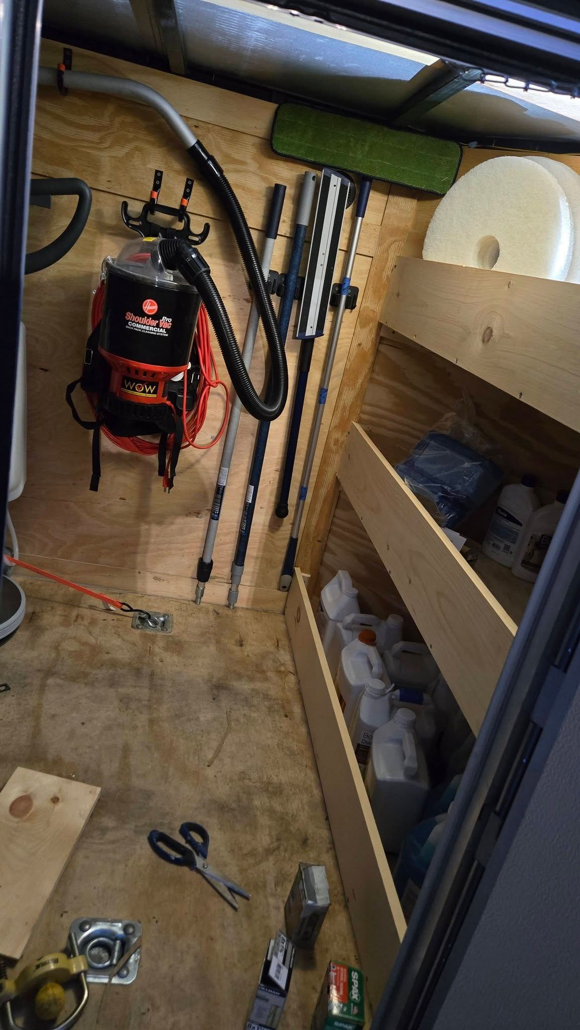 Inside a small shed: red backpack sprayer, fishing rods, shelves, and tools on a plywood floor.