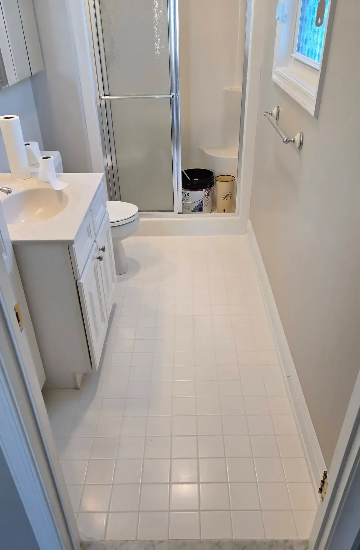 Narrow white bathroom with tiled floor, sink on left, shower at back, and a window on the right.