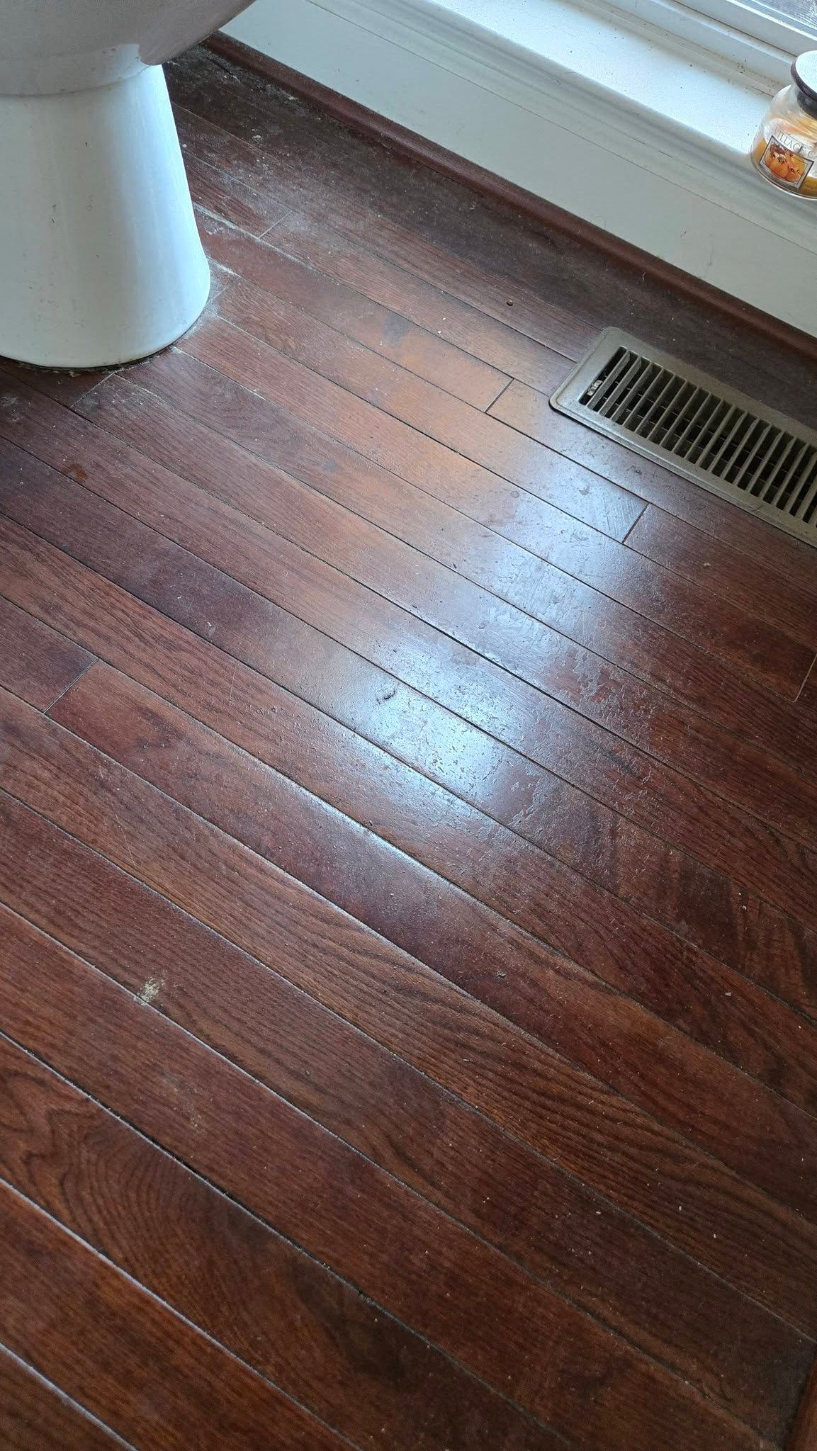 Wooden floorboards beside a white table leg and a floor vent near a window.