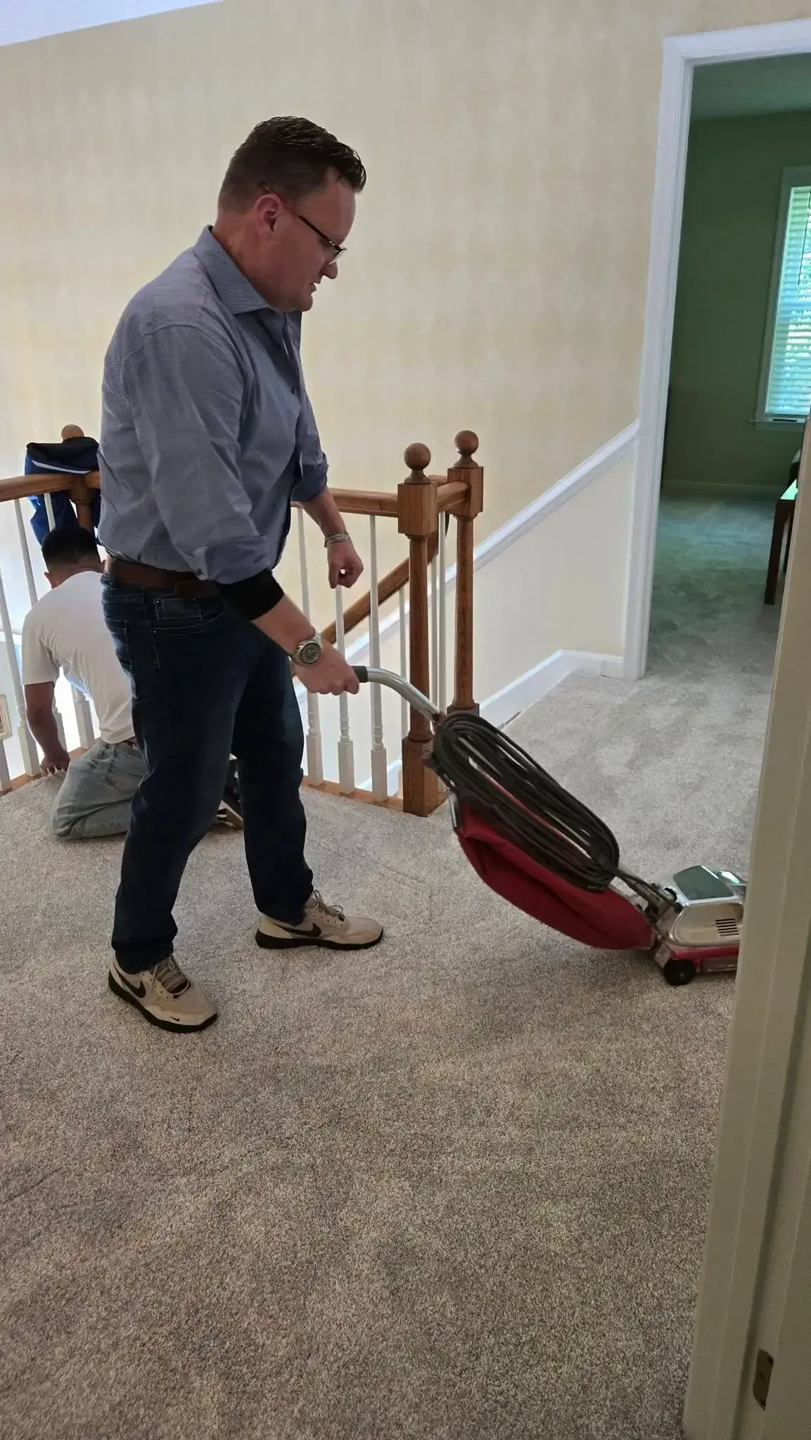 Man vacuuming beige carpet in a hallway near a staircase, with a red upright vacuum.