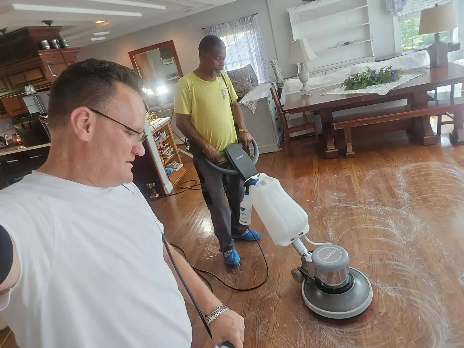 Two men using floor buffers in a sunlit room with hardwood floors