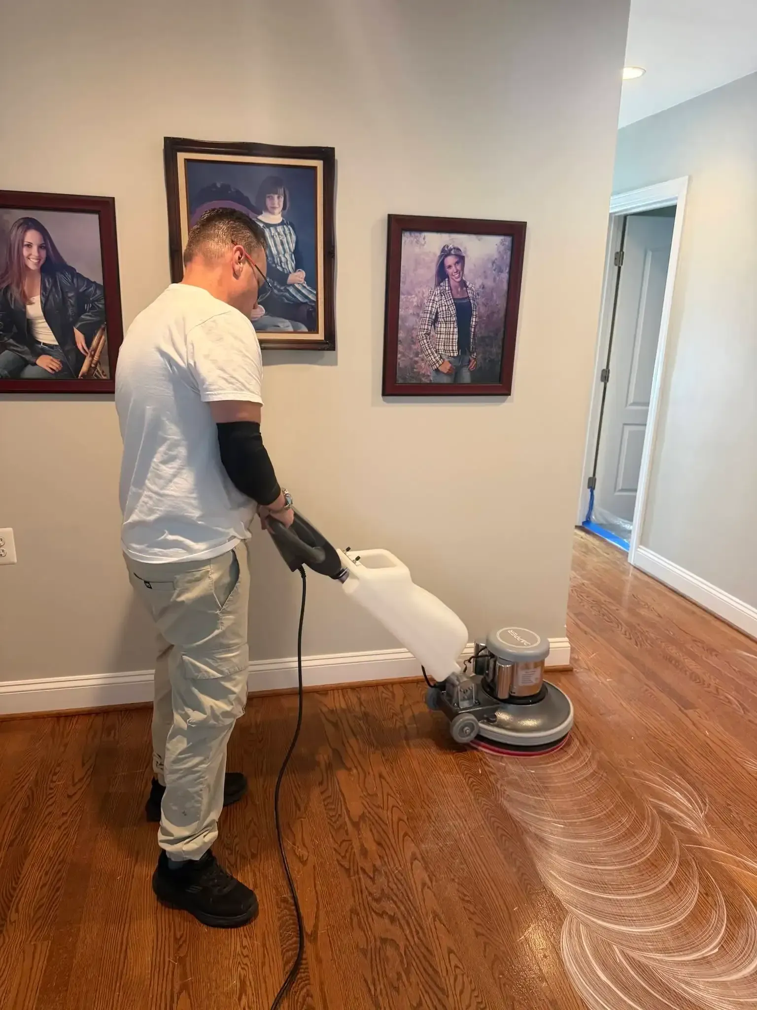 Person polishing a hardwood floor in a hallway with framed portraits on the wall.