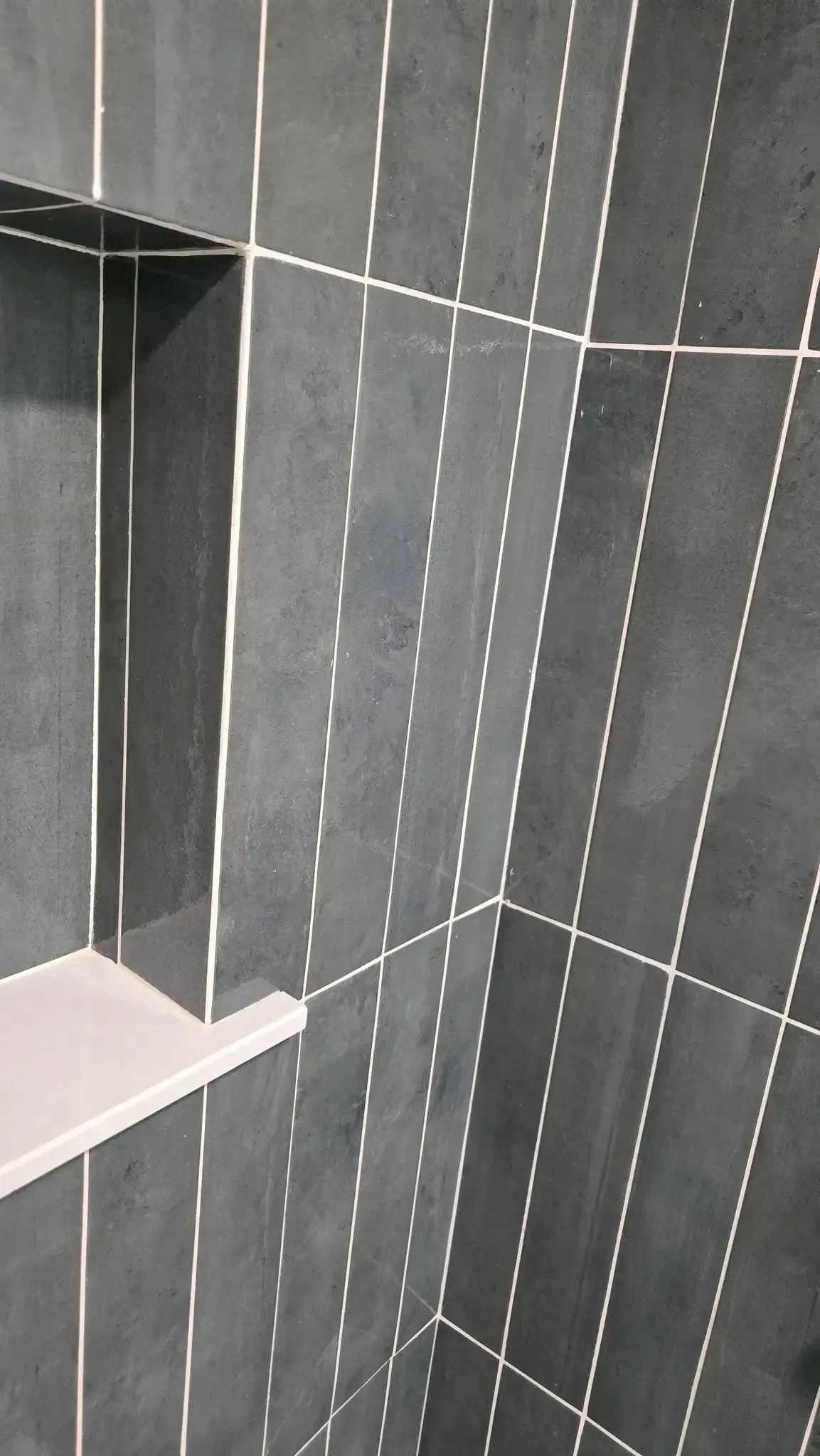 Gray tiled shower corner with a white shelf and a dark doorway on the left
