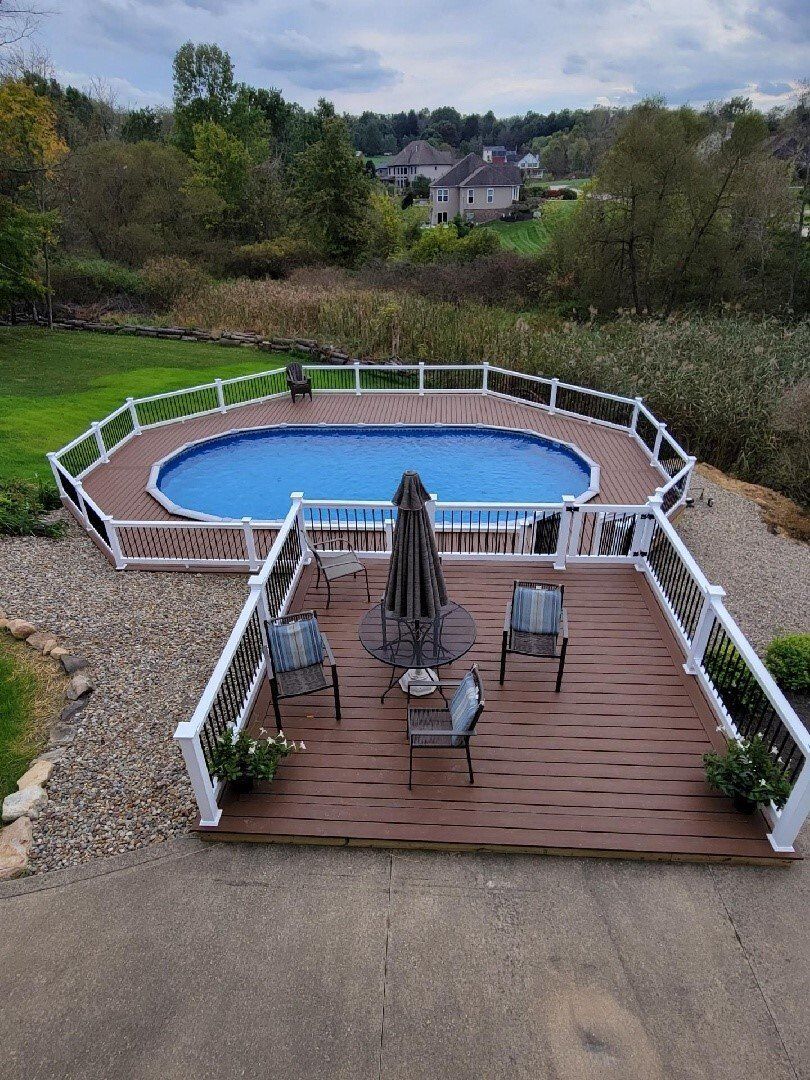 Above-ground oval pool with wooden deck, patio furniture, and umbrella; grassy yard, trees, and houses in the background.