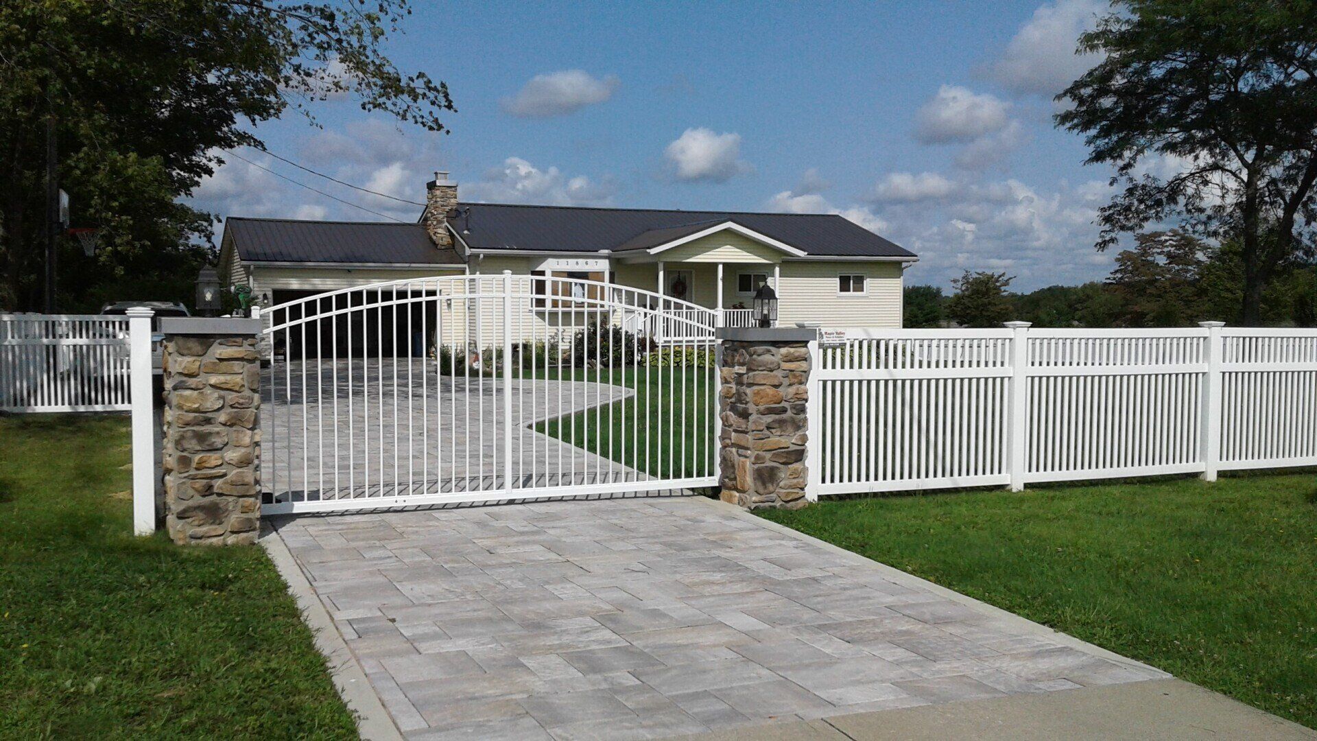 White house with stone gate and driveway, white picket fence, and green lawn under a blue sky.