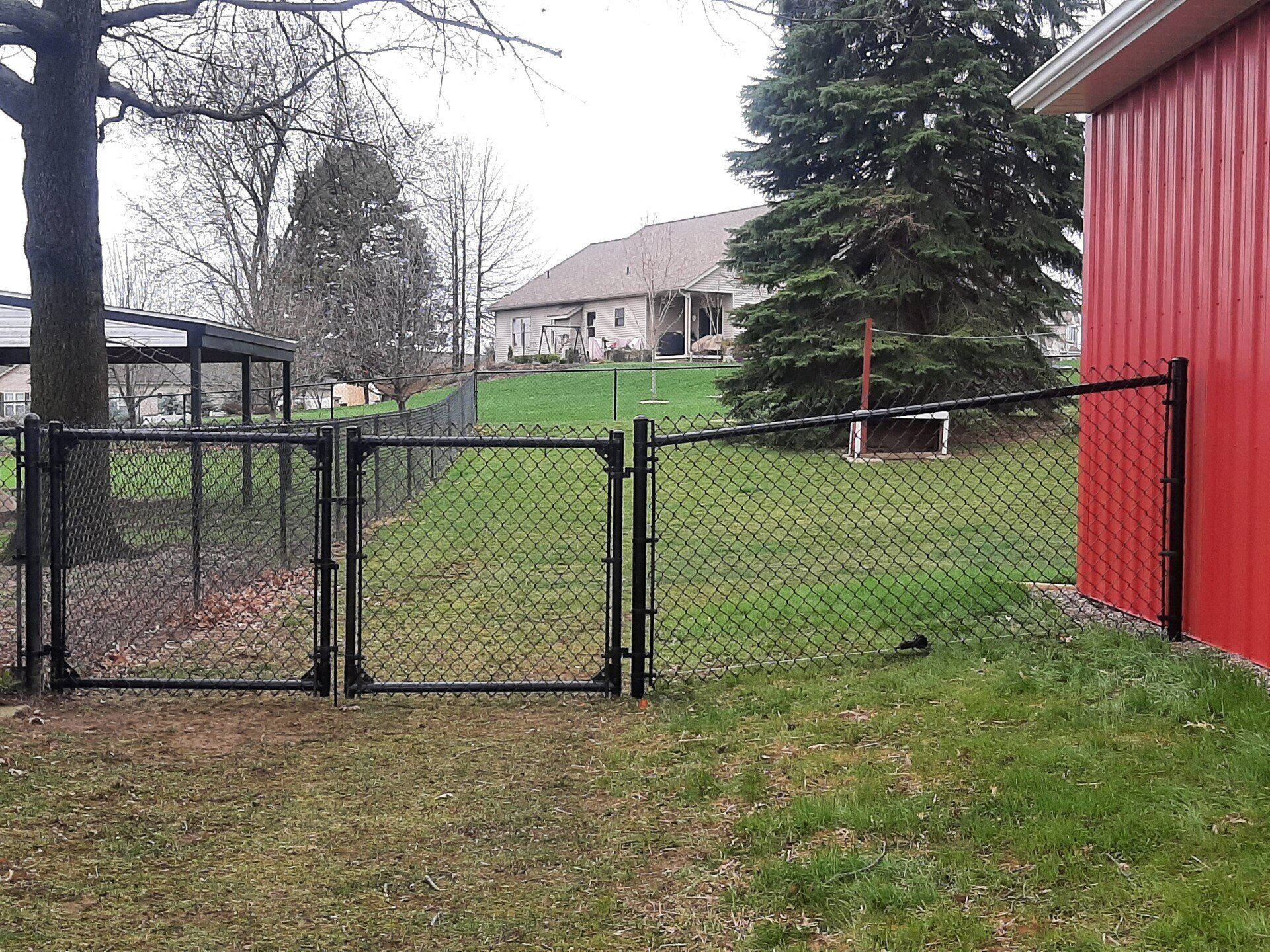 Black chain-link fence with gates beside a red building and grassy yard; a house is in the background.