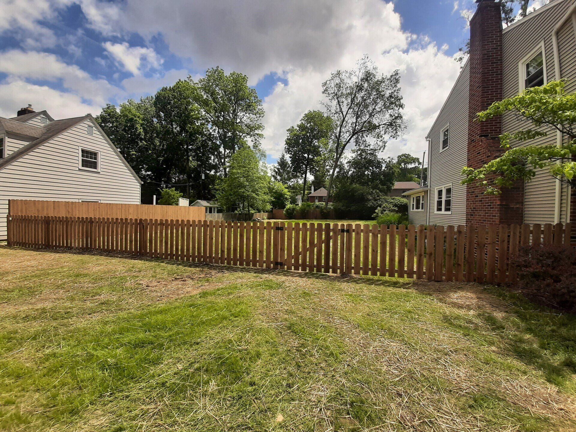 Wooden fence in a grassy yard, between two houses under a cloudy blue sky.