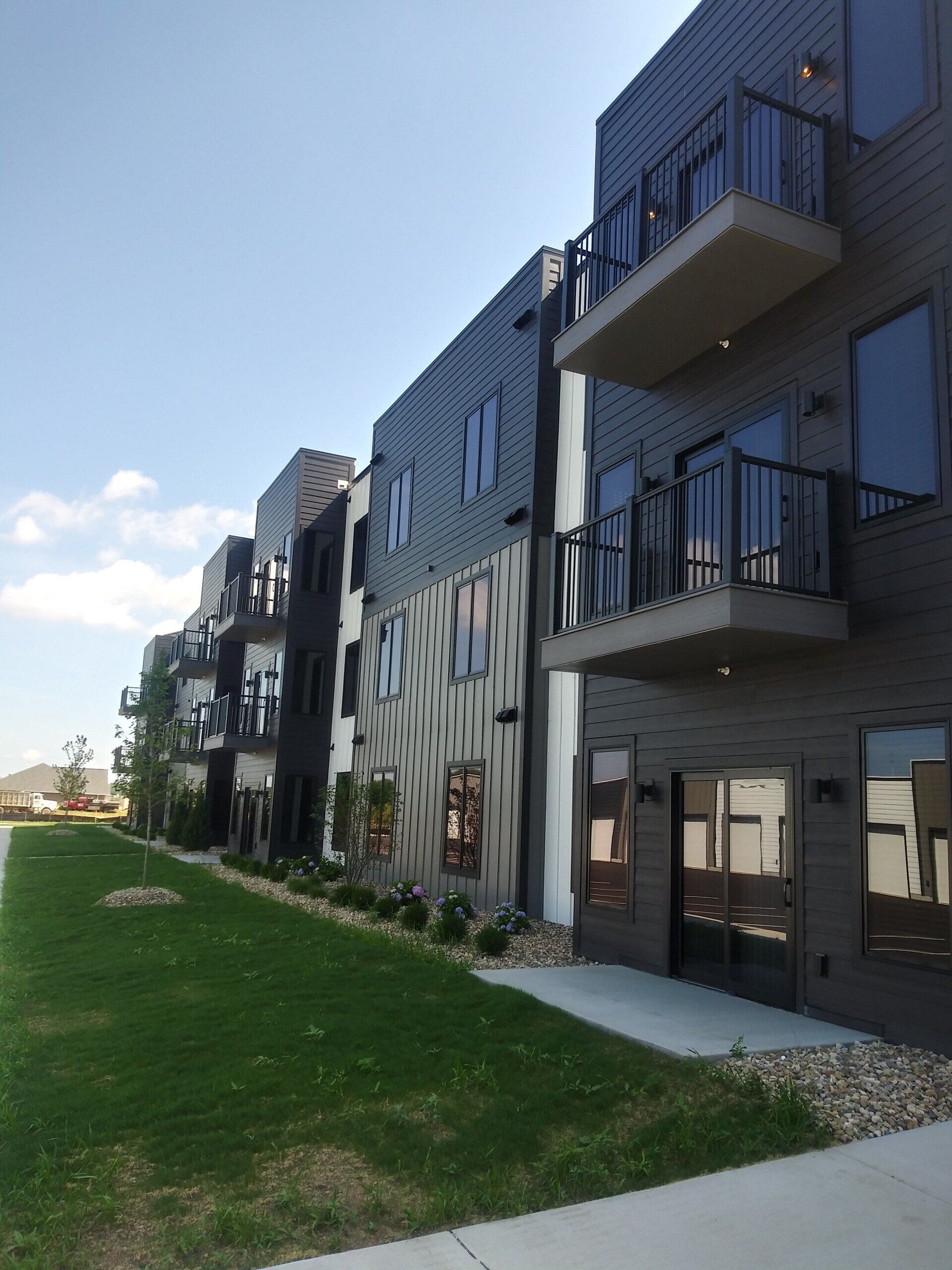 Multi-story modern apartment building with balconies, dark gray siding, and green lawn.