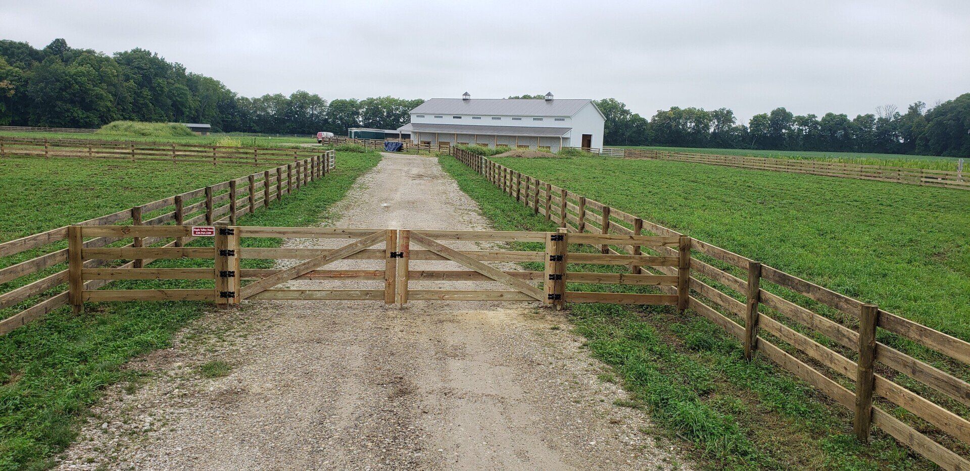 A gravel driveway leads to a white barn flanked by green fields and a wooden fence under an overcast sky.