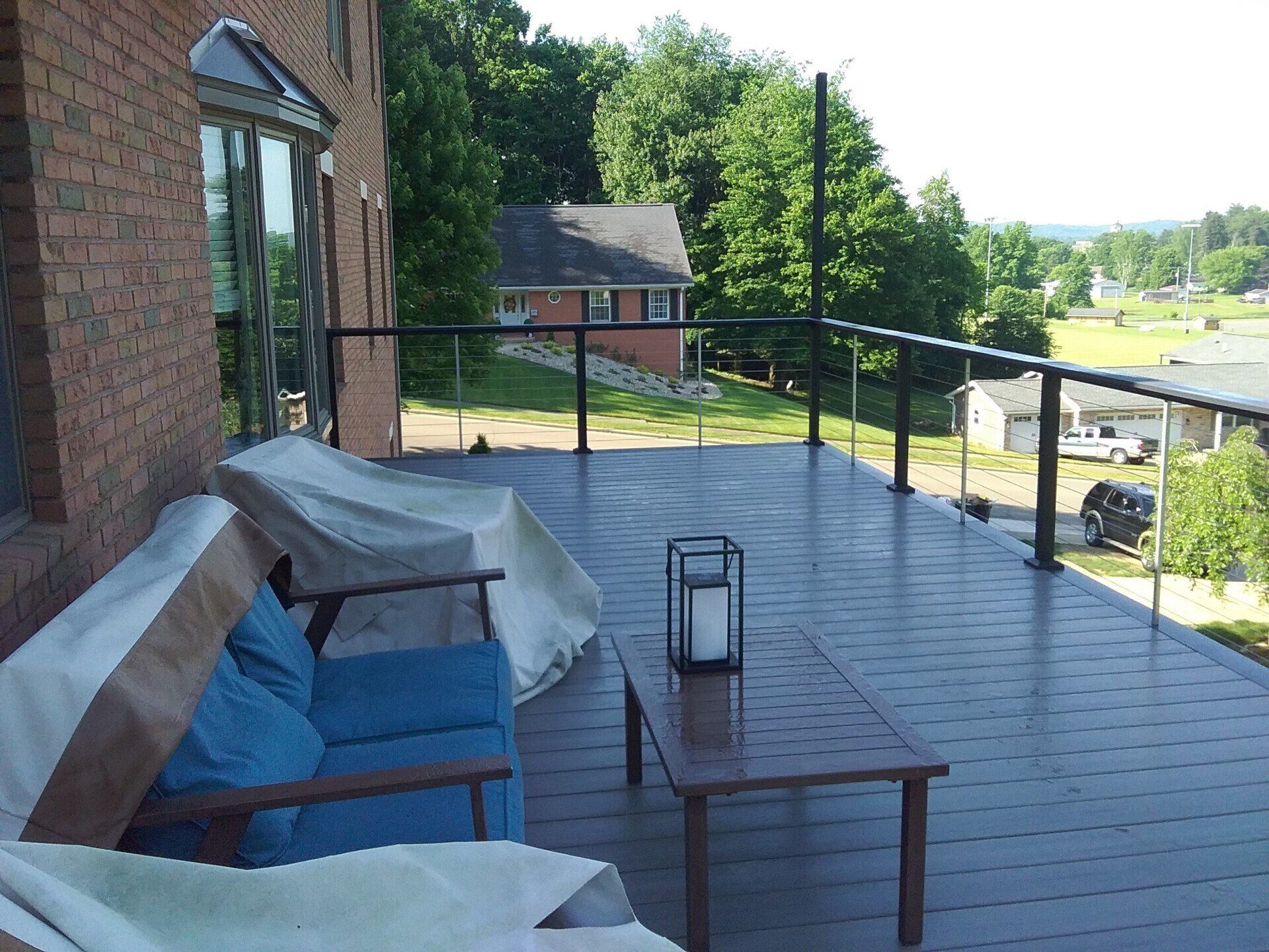 A deck with black railing overlooking a yard. Patio furniture is covered. Brick building. Green trees.