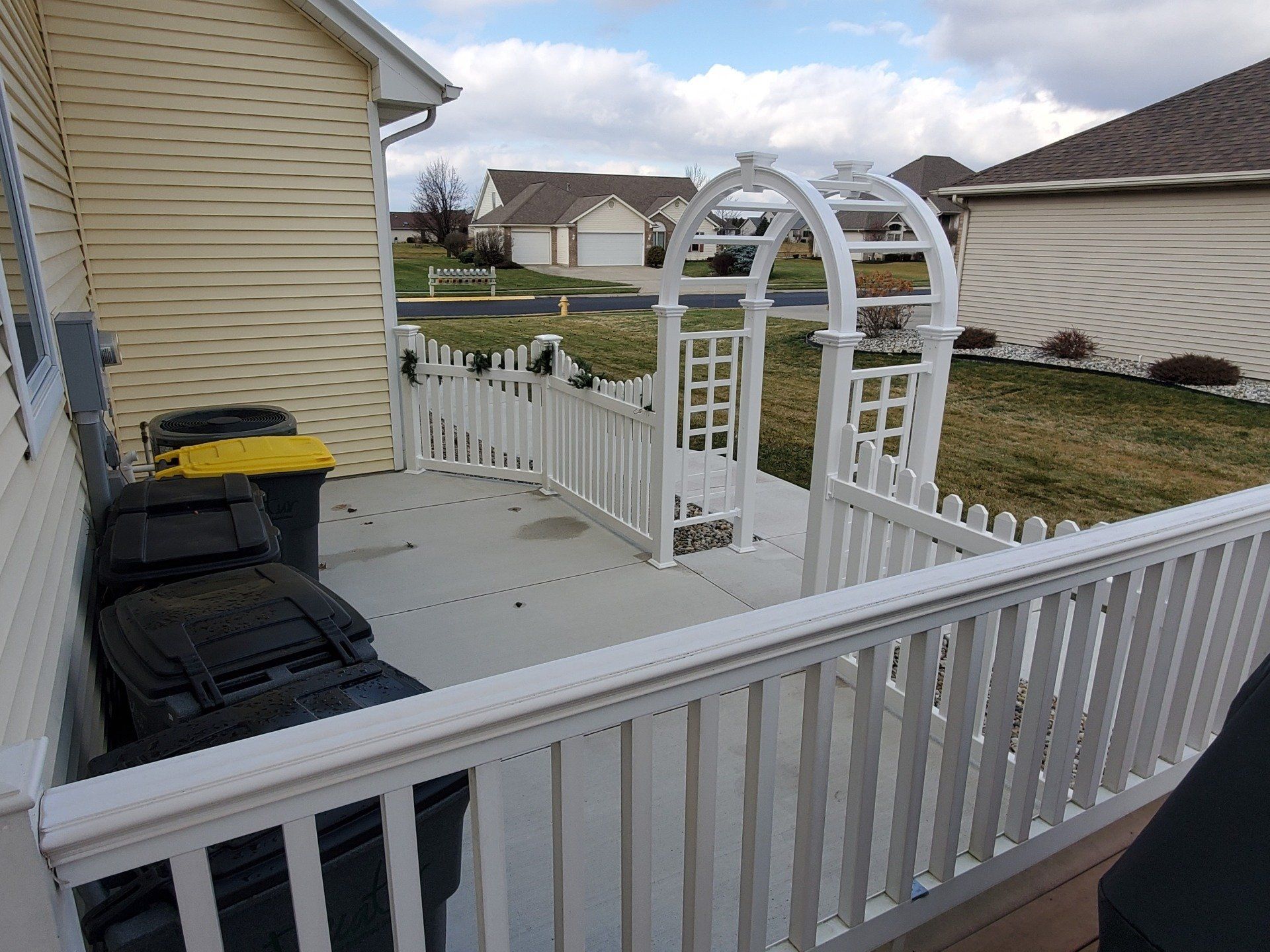 White picket fence with archway on a concrete patio next to a house with black and yellow trash cans.
