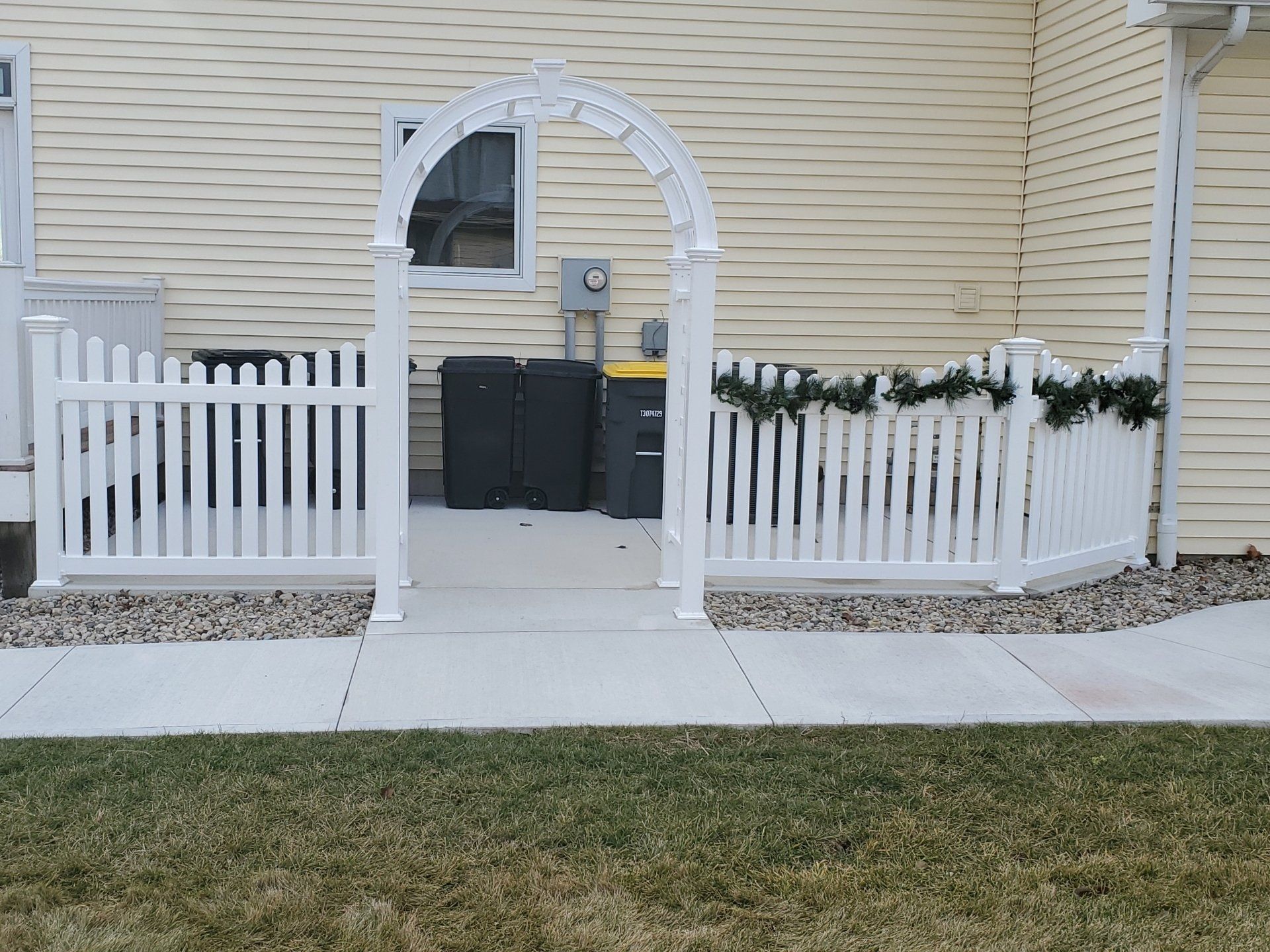 White picket fence with archway entrance on a concrete patio. Green garland decorates the fence.
