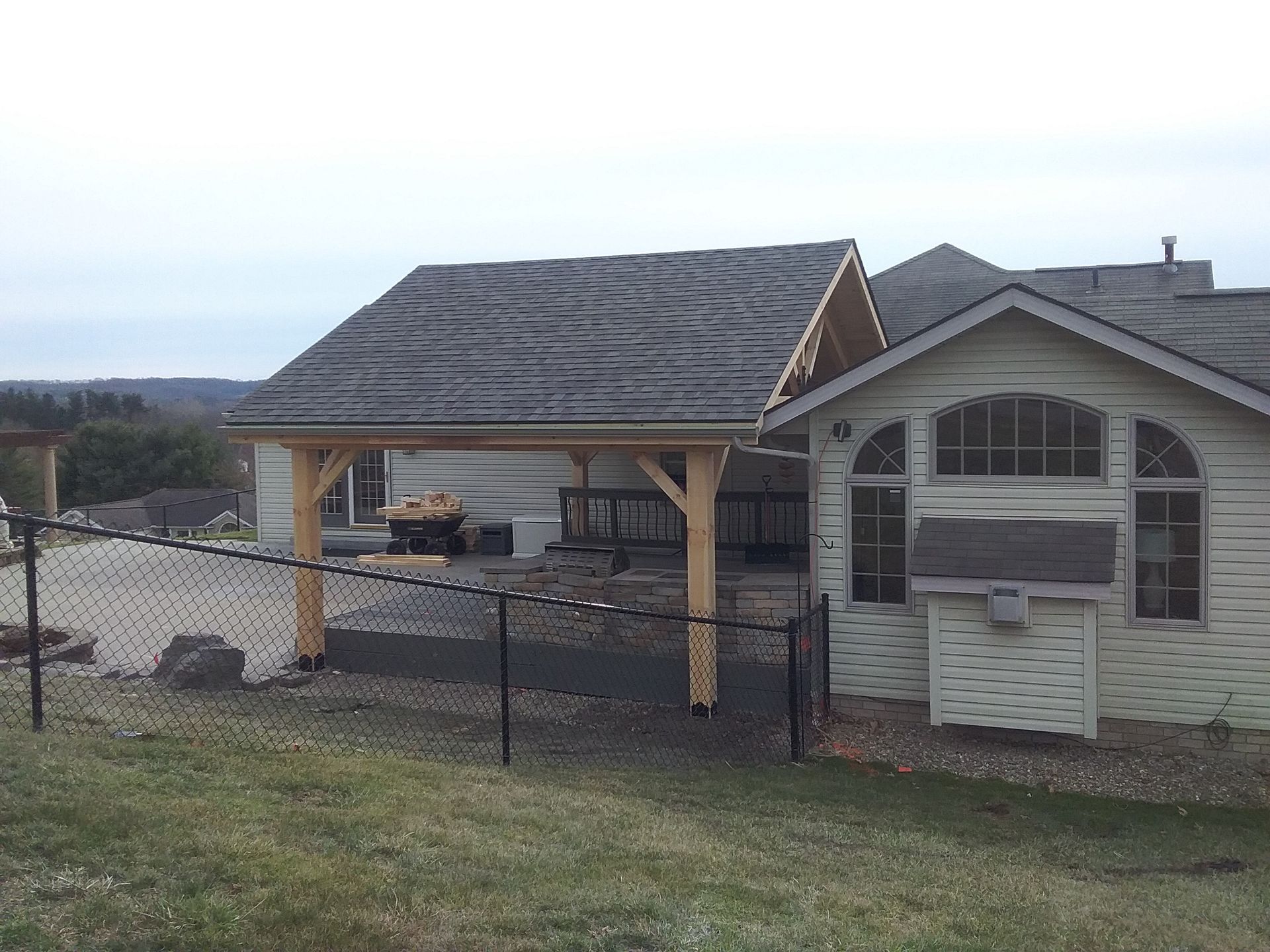 Wooden carport attached to a light-colored house; black chain-link fence in foreground.