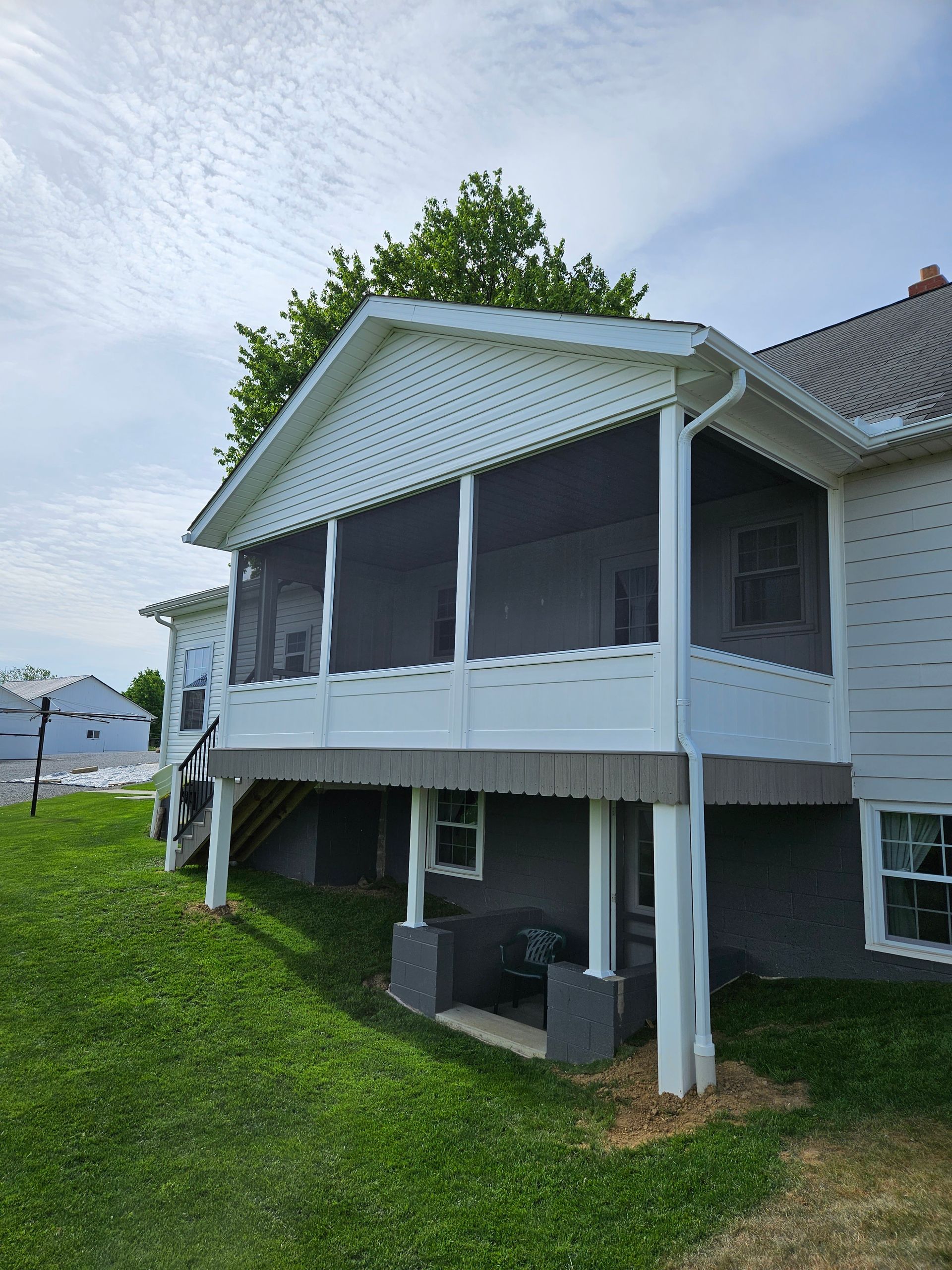 White screened-in porch attached to a white house with grey details, set on a green grassy lawn under a partly cloudy sky.