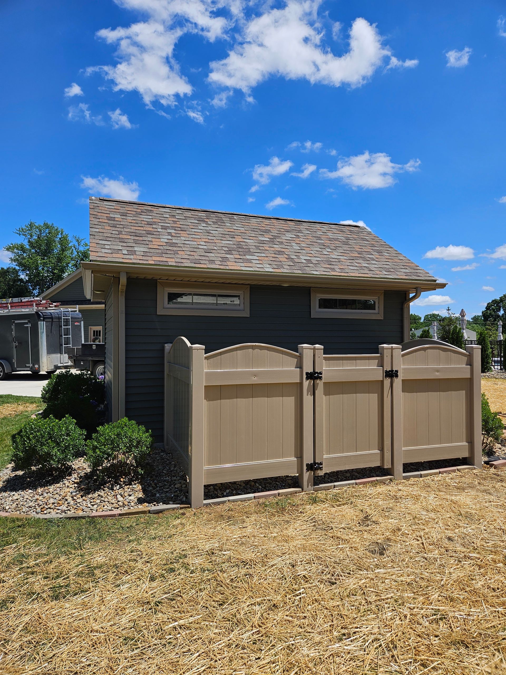 A small, dark gray shed with brown roof, tan windows, and fence under a blue sky.