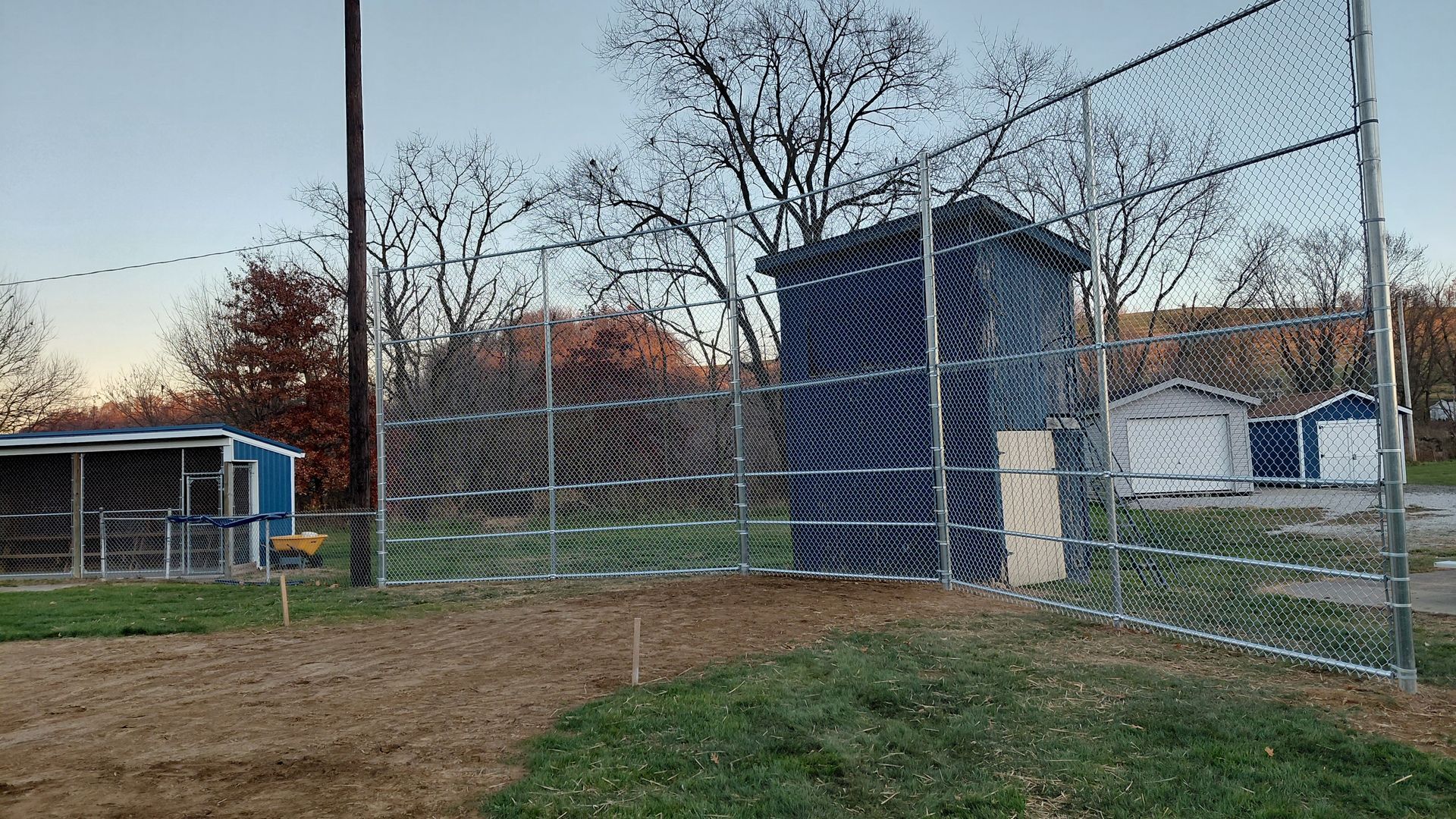 Chain-link fence surrounding a baseball field with a dark blue shed and blue buildings in the background.