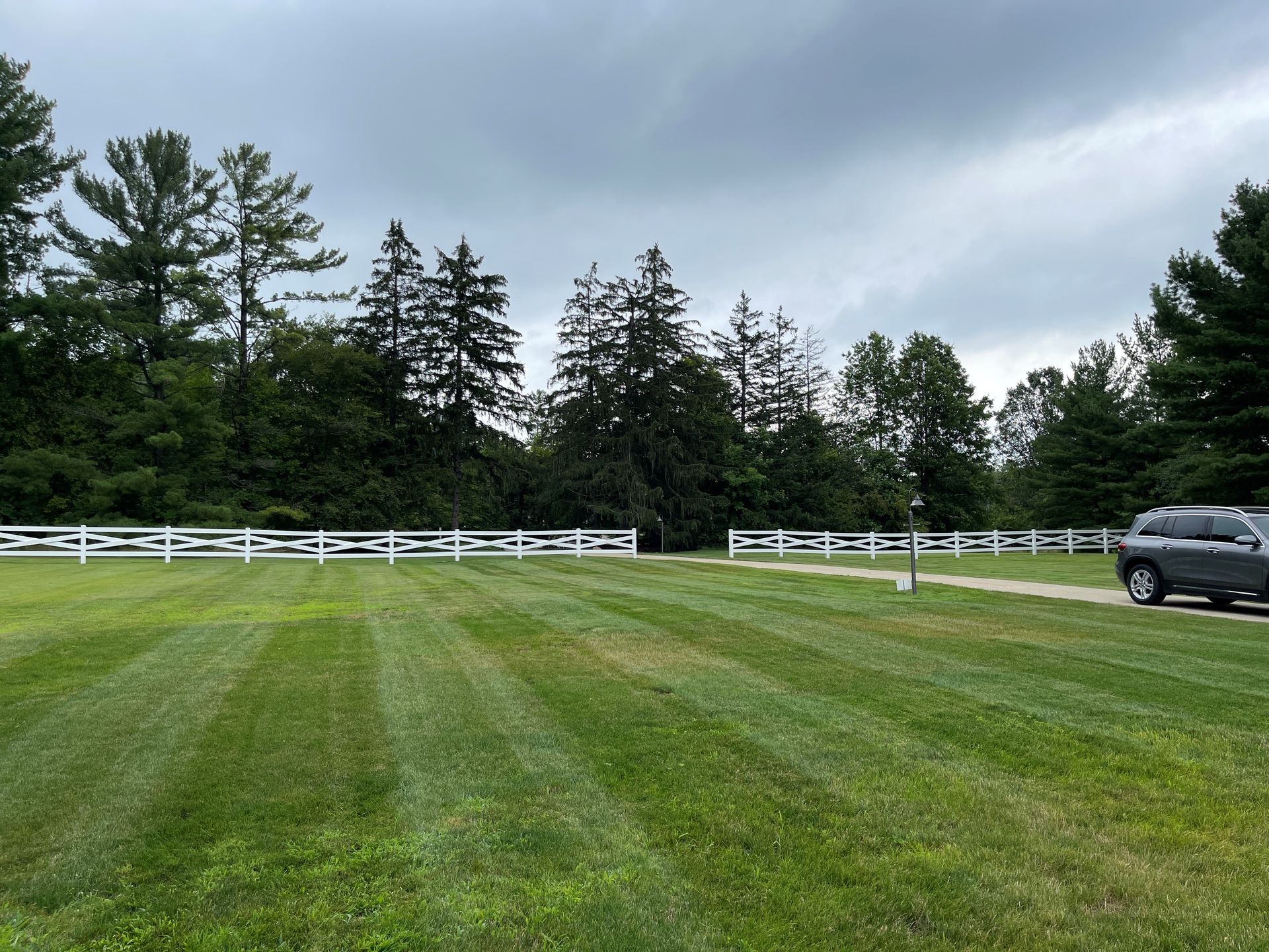 Lawn in front of white fence, trees and cloudy sky. A gray SUV is parked on the right.