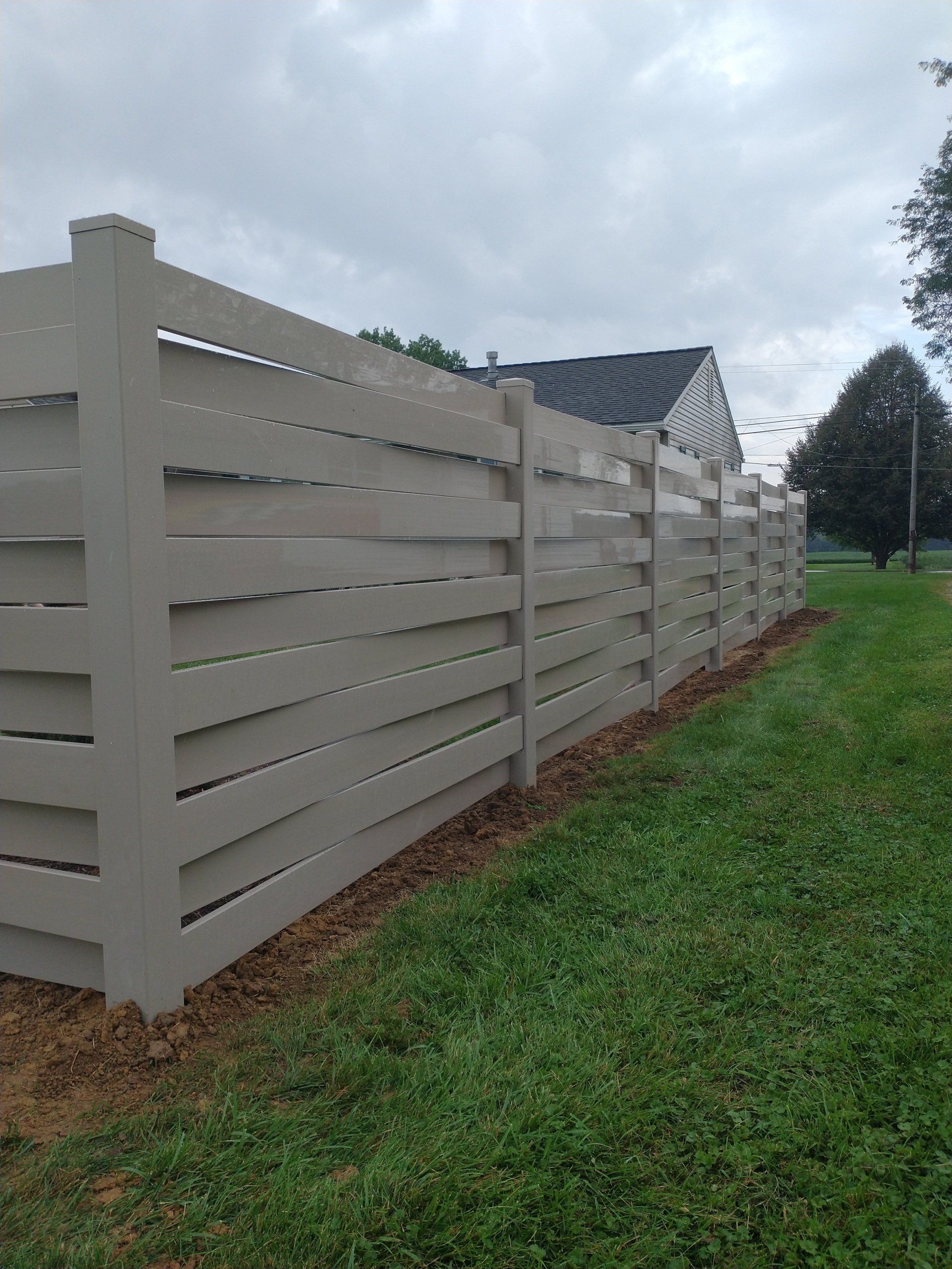 A tan horizontal slat fence along a grassy lawn, under a cloudy sky.