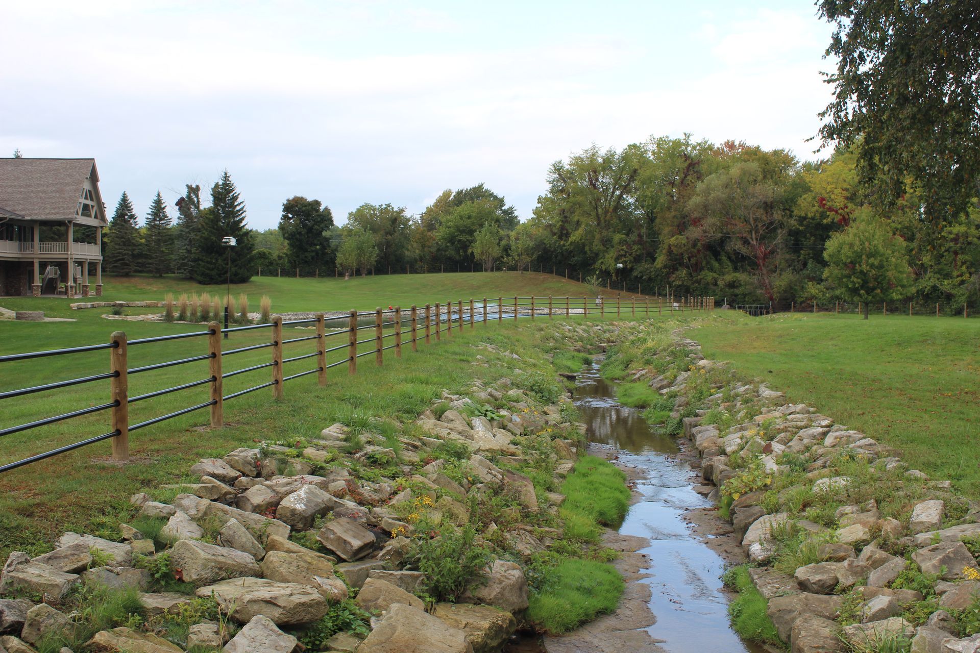 Creek flowing through a grassy area, bordered by rocks and a fence, with trees and a building in the background.