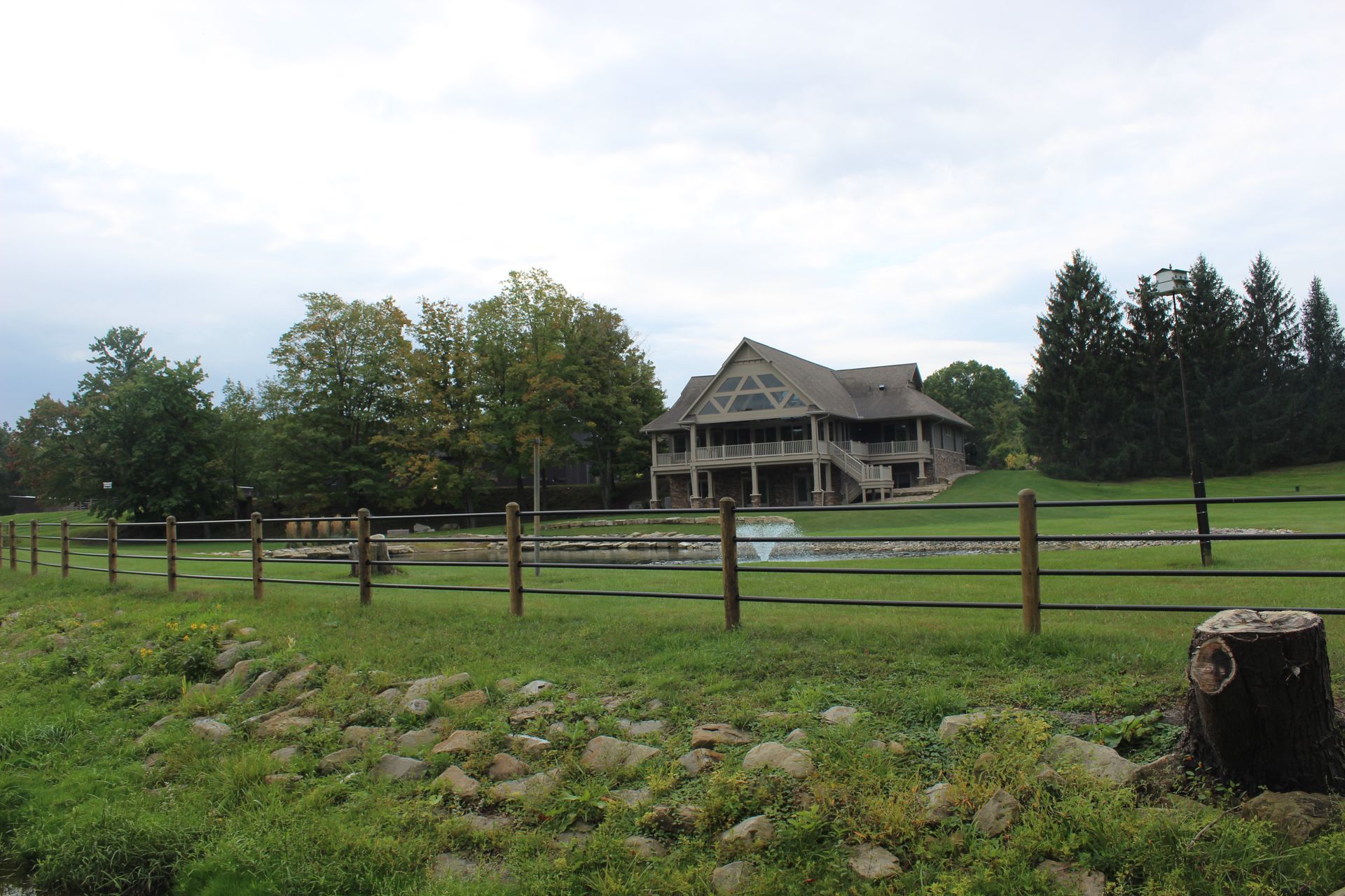 Large wooden house with multiple levels, surrounded by a fence and green field, trees in background, overcast sky.