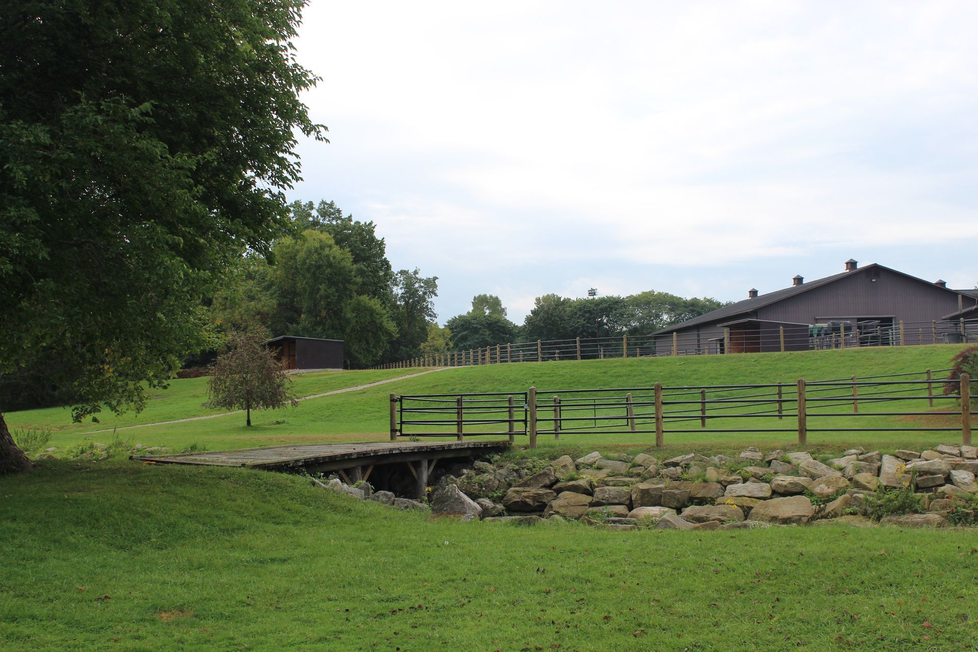 Green field with small bridge over a creek. Wooden fence and buildings under a cloudy sky.
