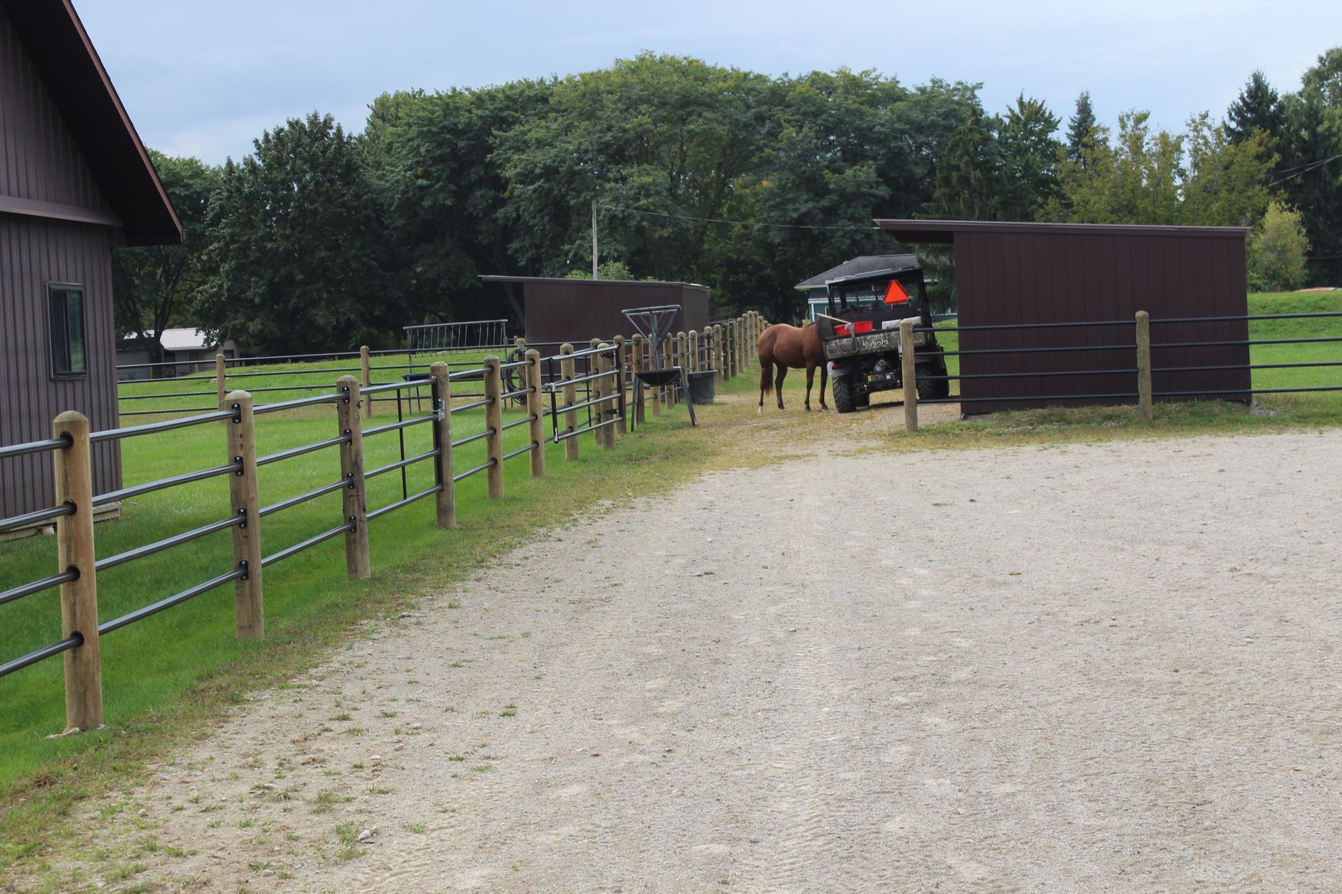 A gravel driveway leads to a stable with horses. Brown buildings and fences are visible.