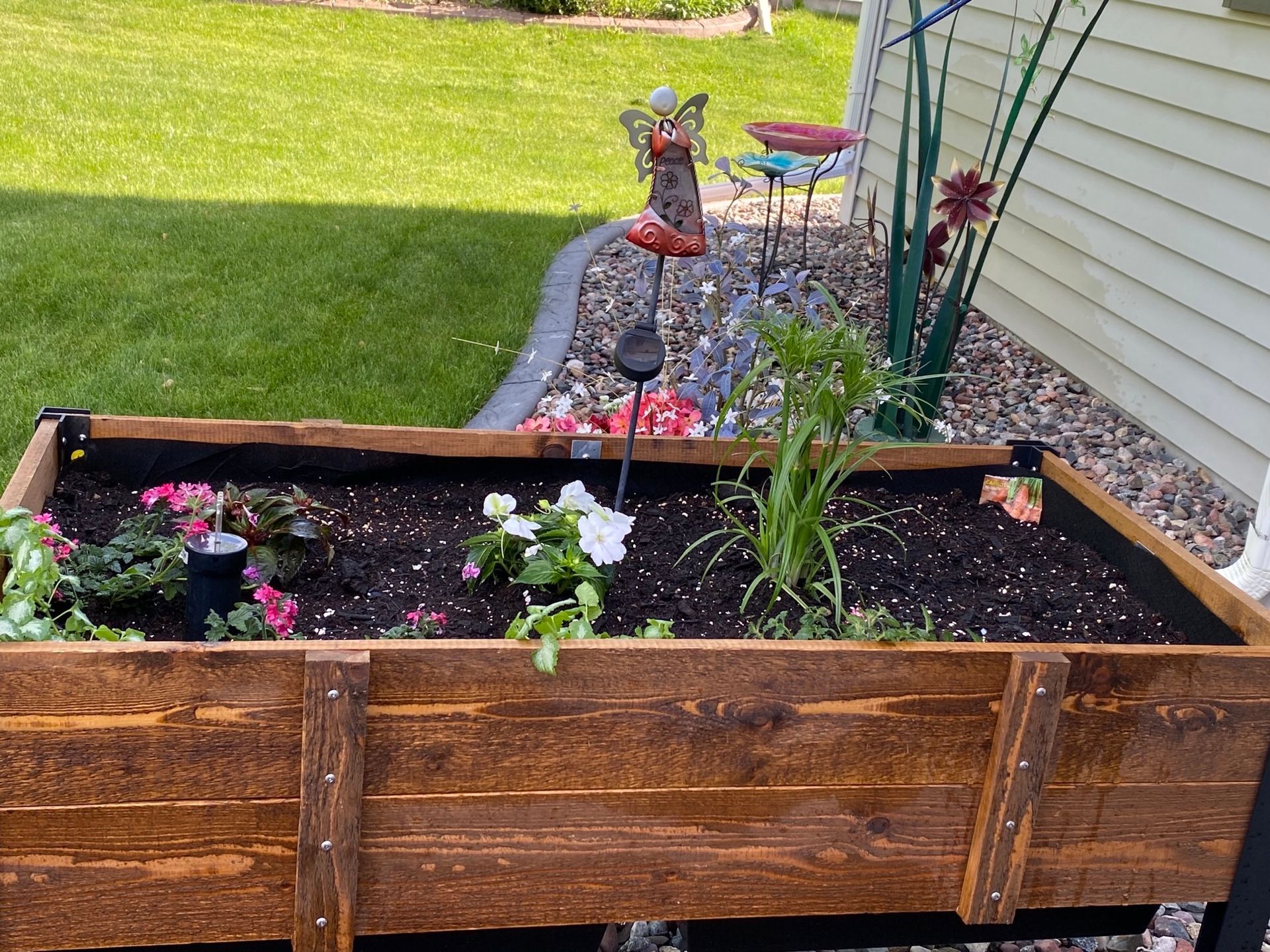 Wooden raised garden bed with flowers and garden art. Grassy lawn and house siding in the background.